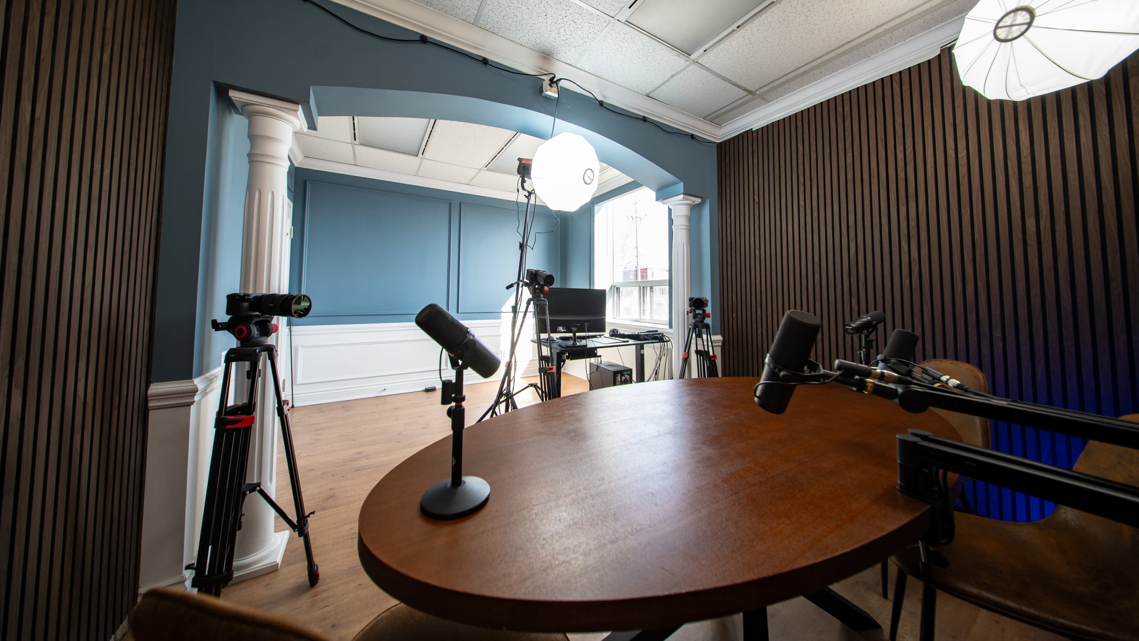 A recording studio is set up with a round wooden table, several microphones on stands, camera tripods, and a backdrop featuring wooden paneling.