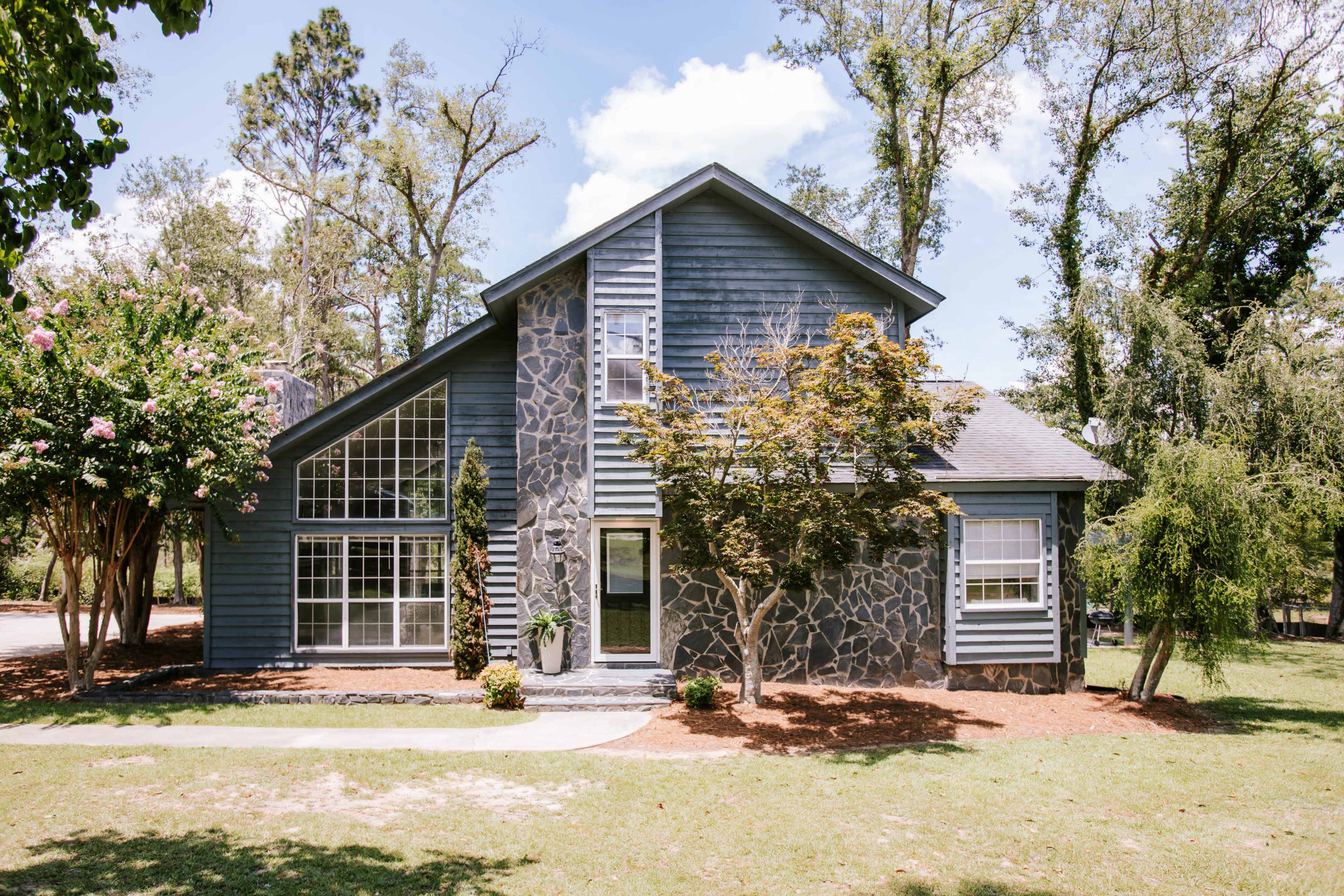 The image shows a modern two-story house with a stone and blue wood exterior, surrounded by green trees and landscaping.