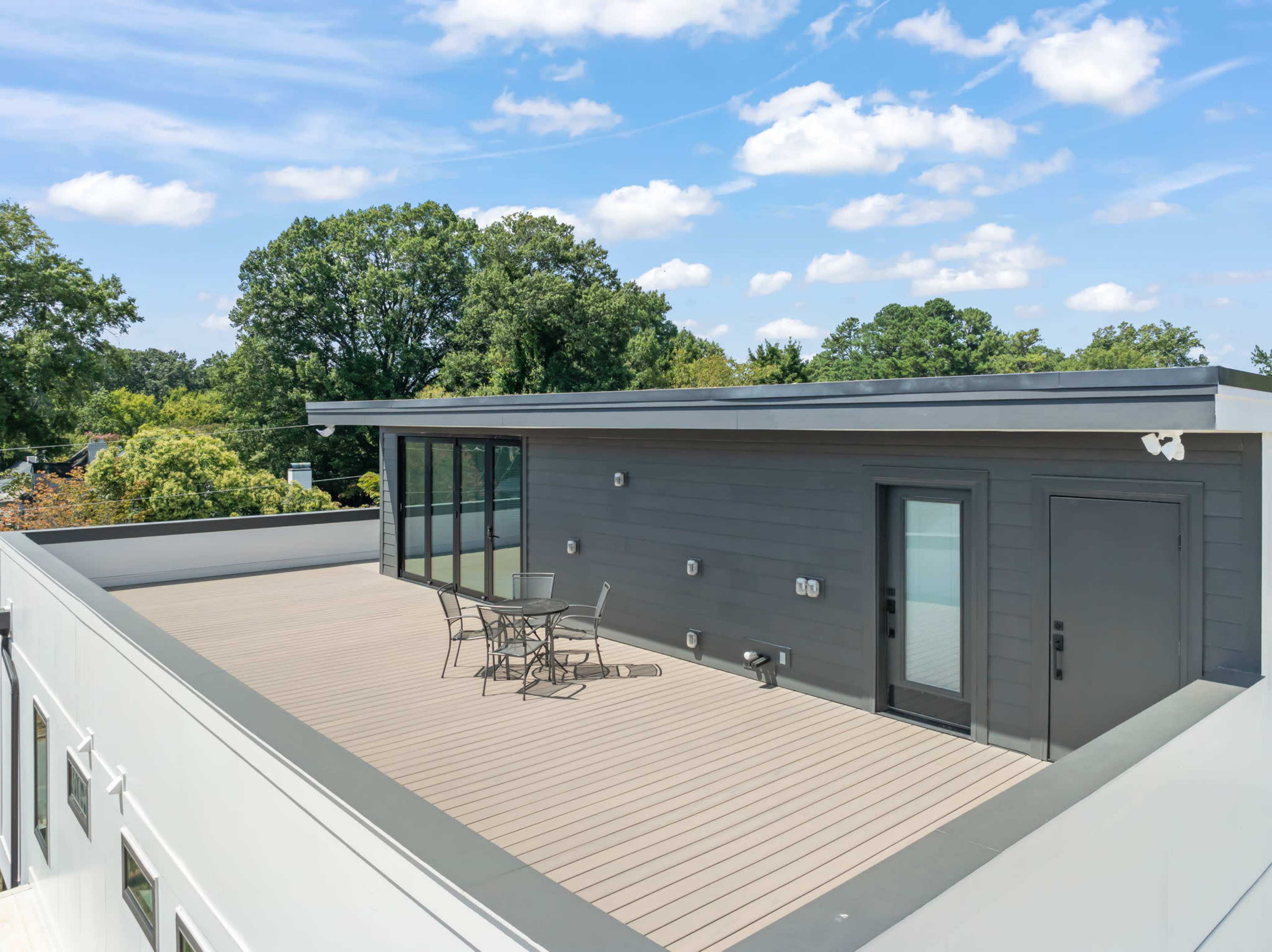 A flat rooftop deck features a small table and chairs, surrounded by greenery and under a blue sky with scattered clouds.
