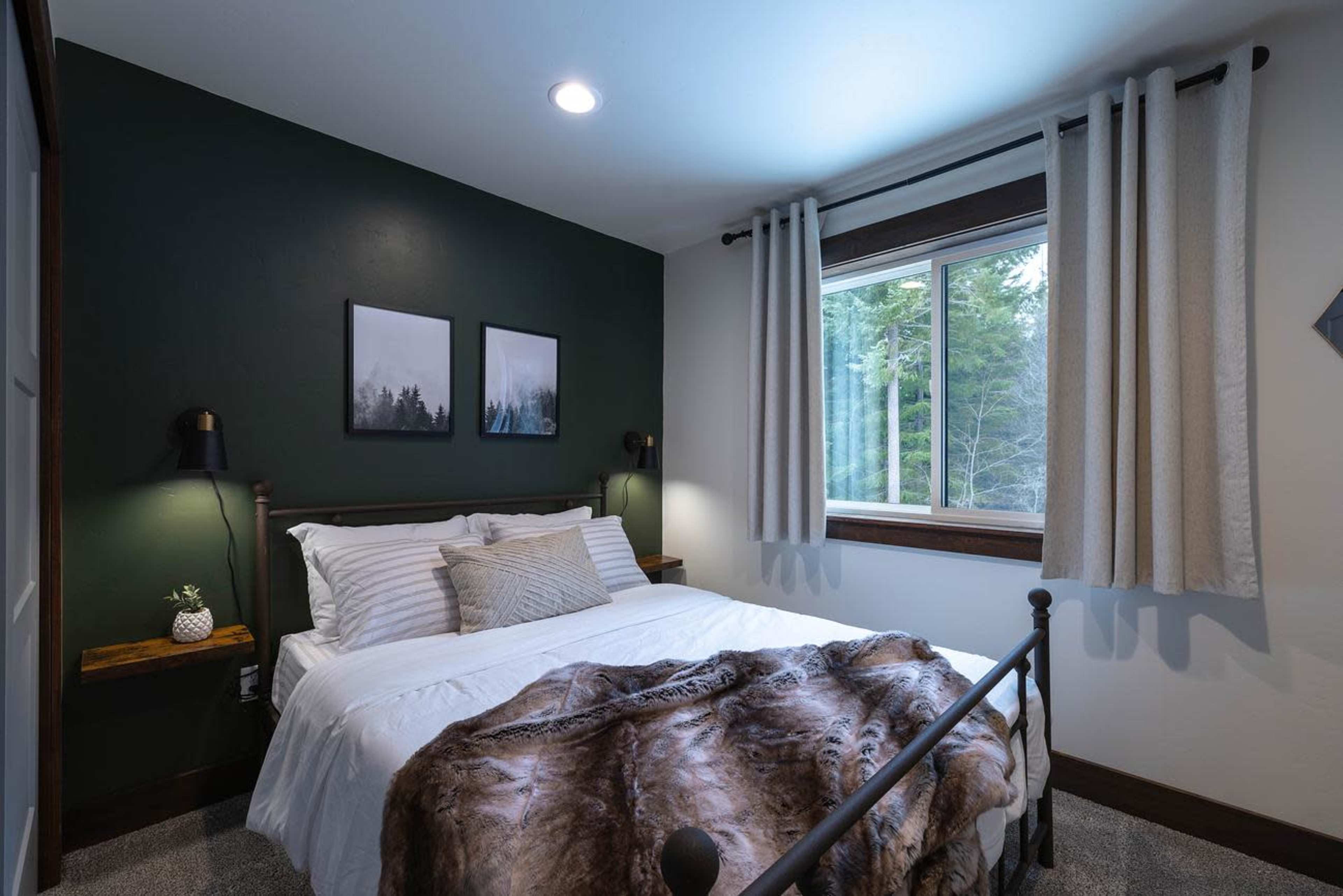 A bedroom with a bed, two framed pictures on the wall, and a window revealing a view of trees outside.