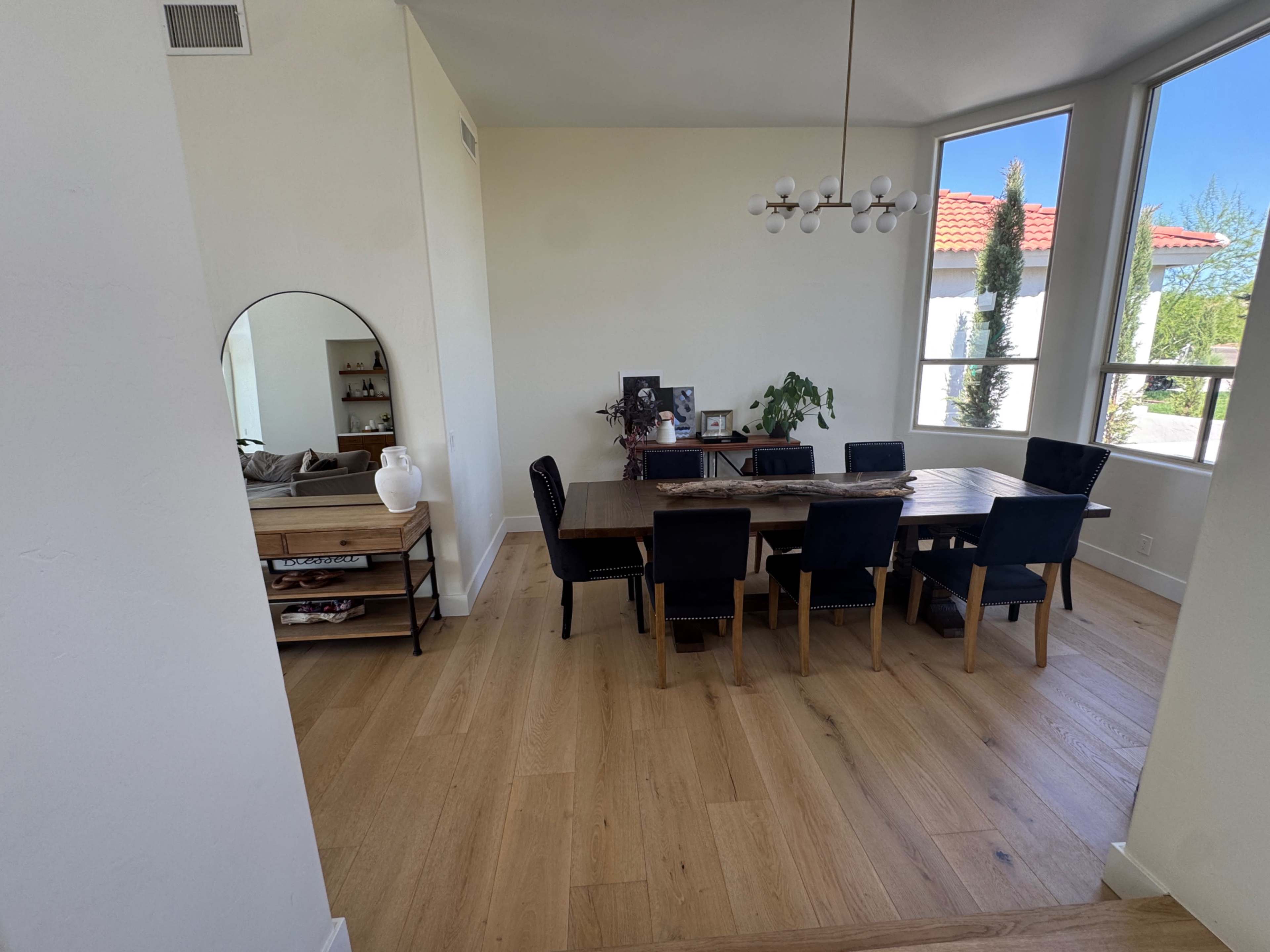 The image shows a modern dining area with a wooden table surrounded by black chairs, a large mirror on the wall, and a view of greenery through large windows.