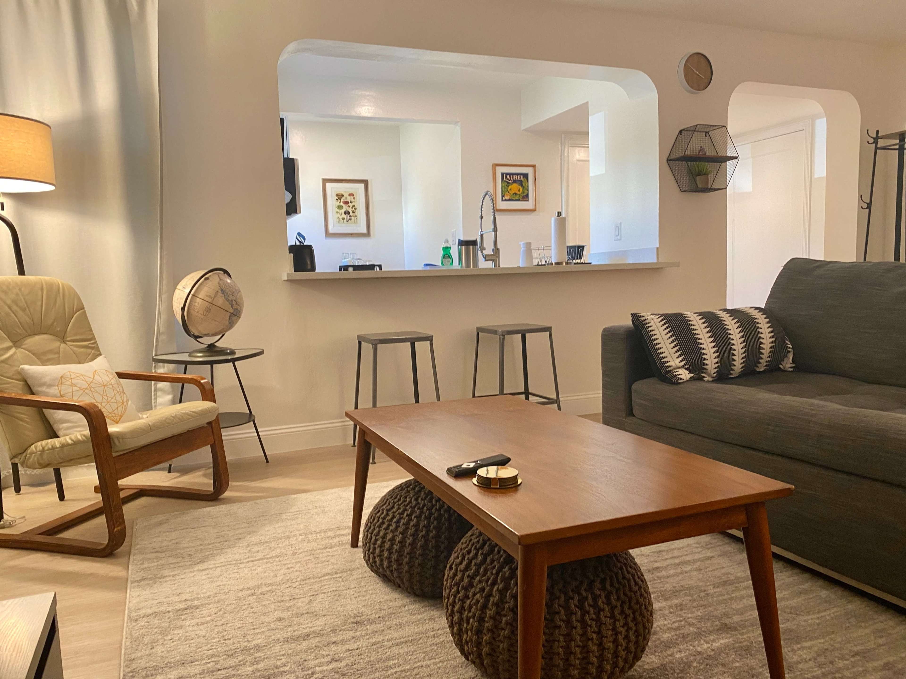 A modern living room with a wooden coffee table, a gray sofa, a beige armchair, and a kitchen area visible through an archway.