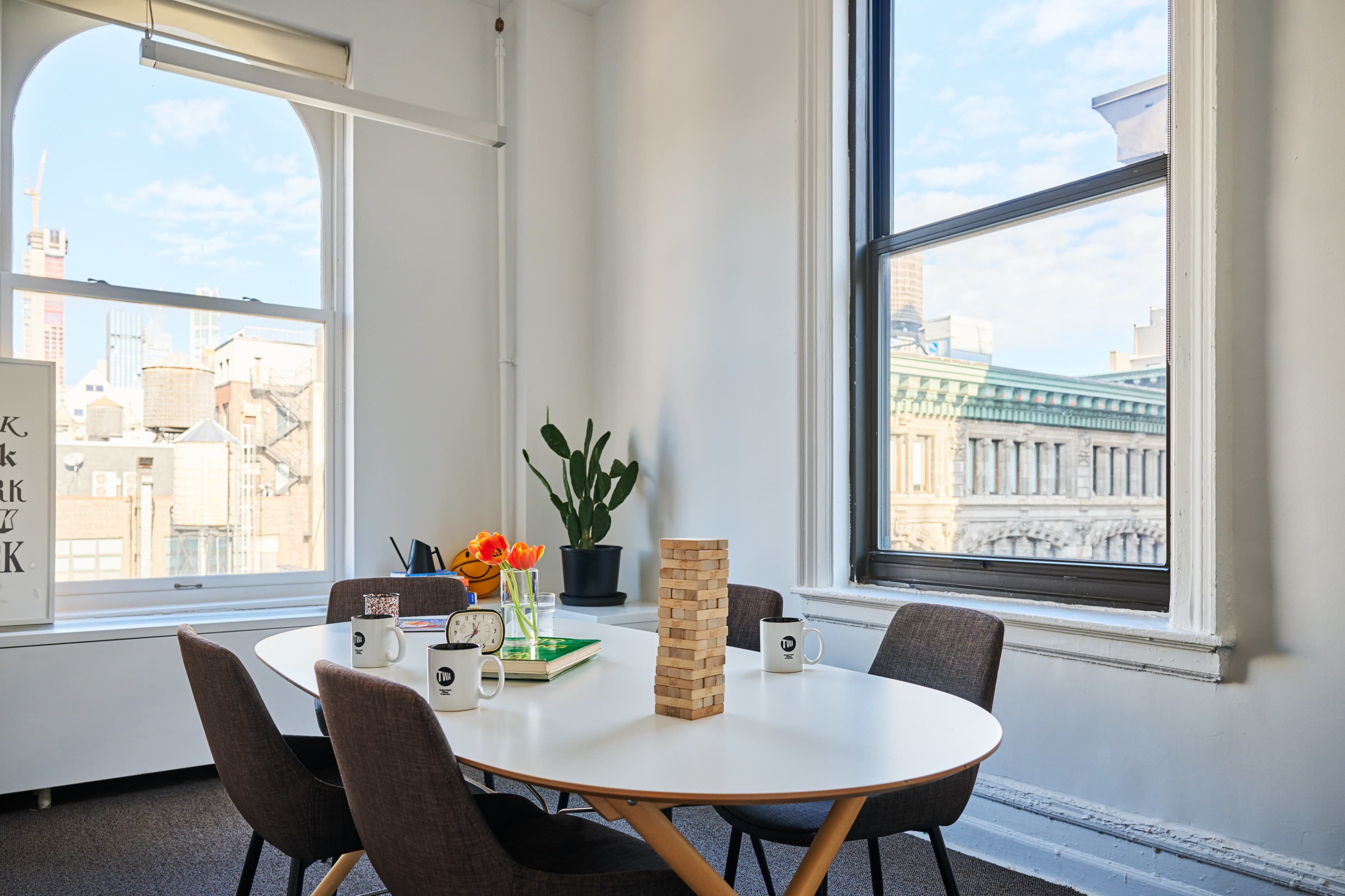 A bright meeting room features a white oval table surrounded by chairs, a plant by the window, and a Jenga tower as a centerpiece.