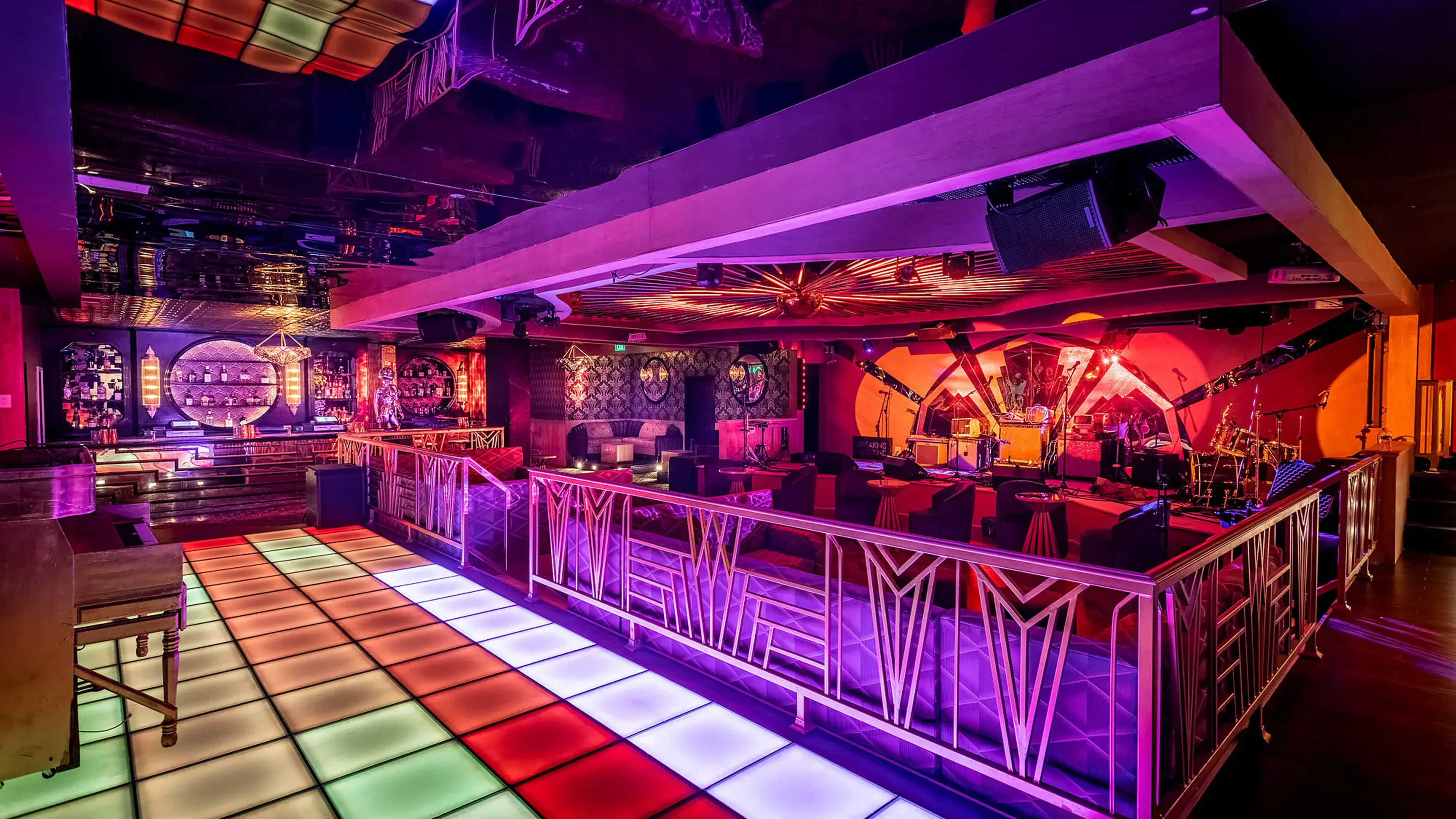 The image shows a vibrant nightclub interior, featuring a colorful dance floor with illuminated tiles and a bar area in the background, set against a backdrop of artistic lighting and decor.
