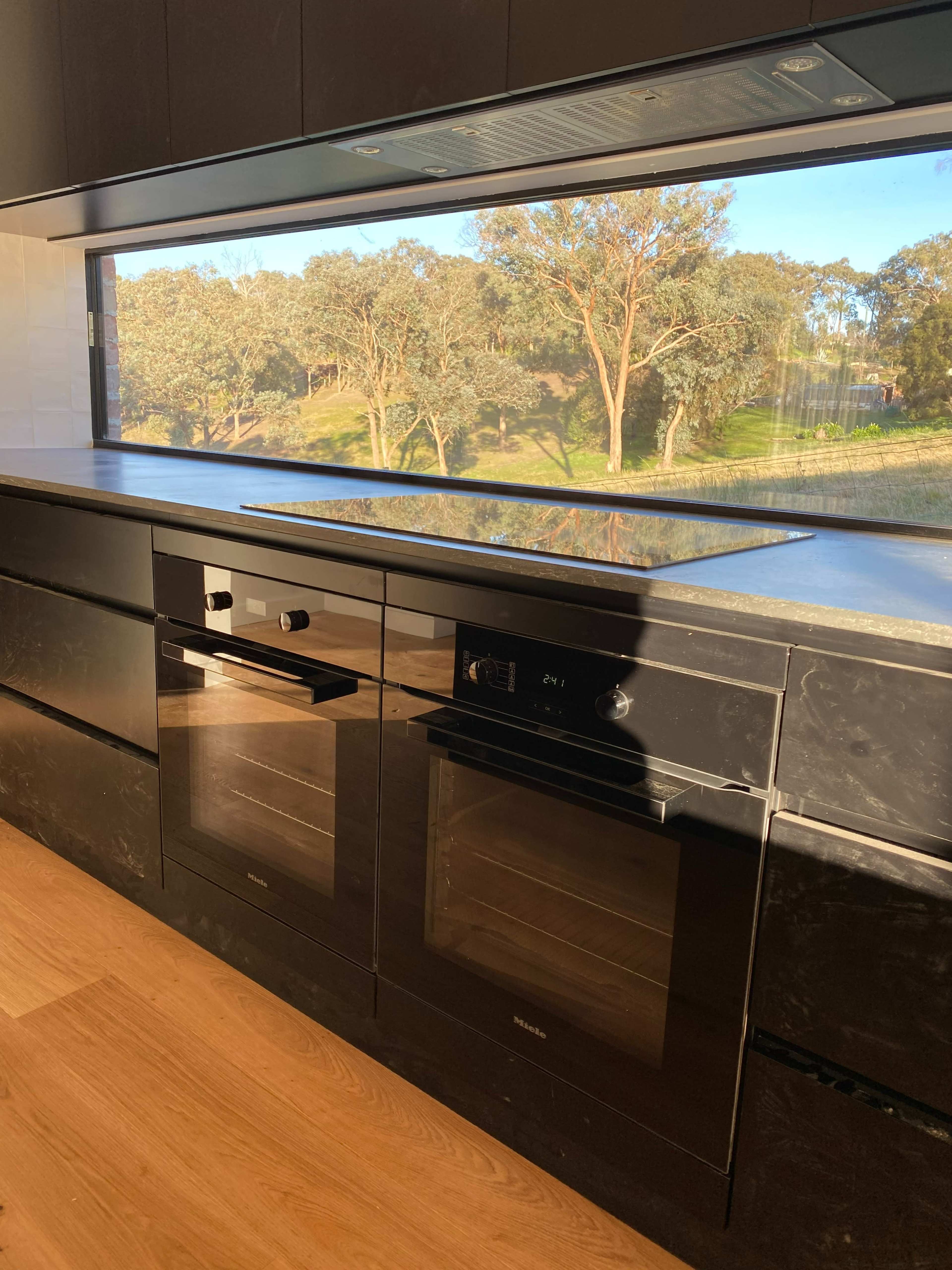 A modern kitchen with black cabinetry, built-in ovens, and a large window overlooking trees and greenery outside.