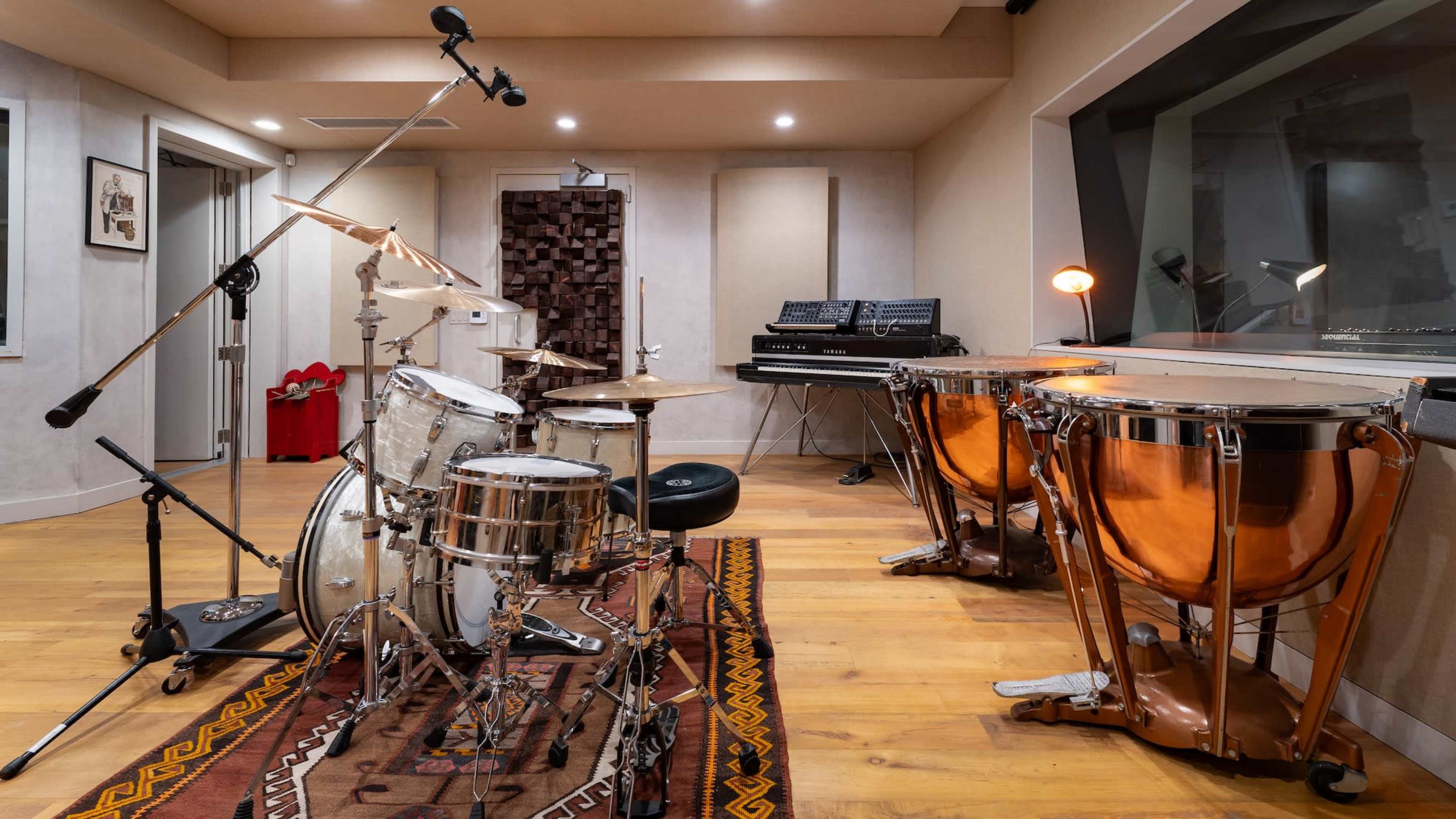 The image shows a music recording studio featuring a drum set, a pair of conga drums, and various audio equipment arranged in a well-lit room with wooden flooring.