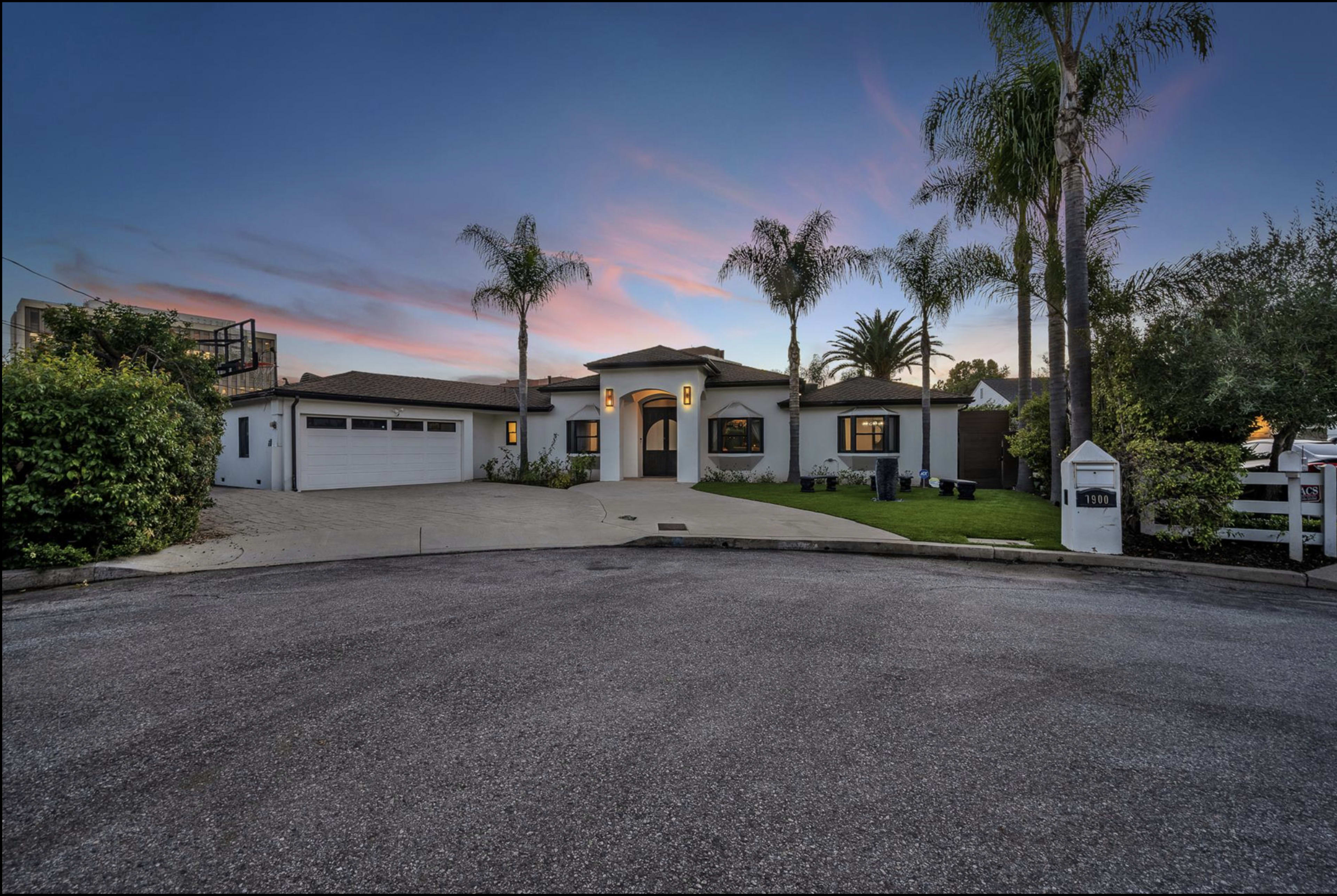 A modern single-story house with palm trees and a landscaped front yard sits at the end of a cul-de-sac under a colorful sunset sky.