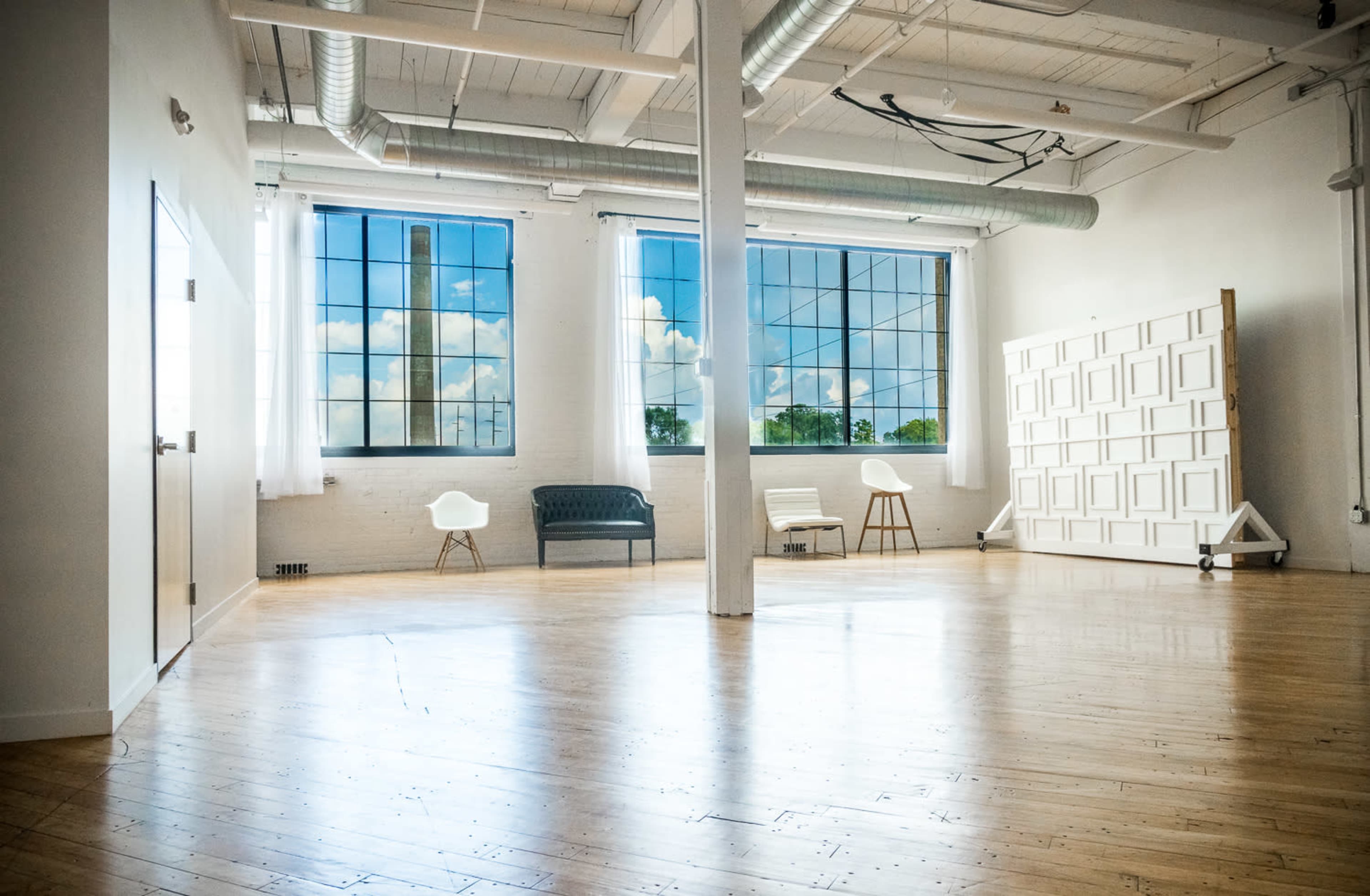 A spacious, bright room with wood flooring, large windows, and minimal furniture including a gray sofa and white chairs.