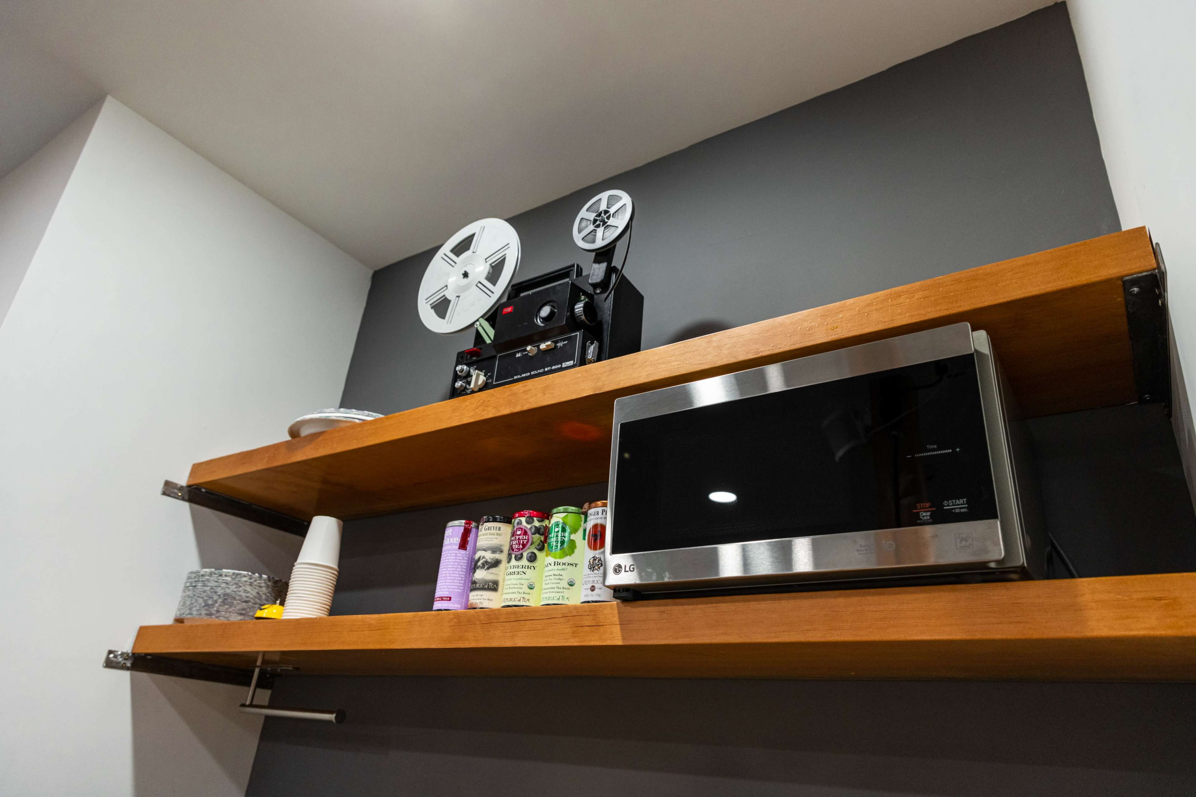 A black and silver microwave and a vintage film projector placed on wooden shelves against a dark grey wall.