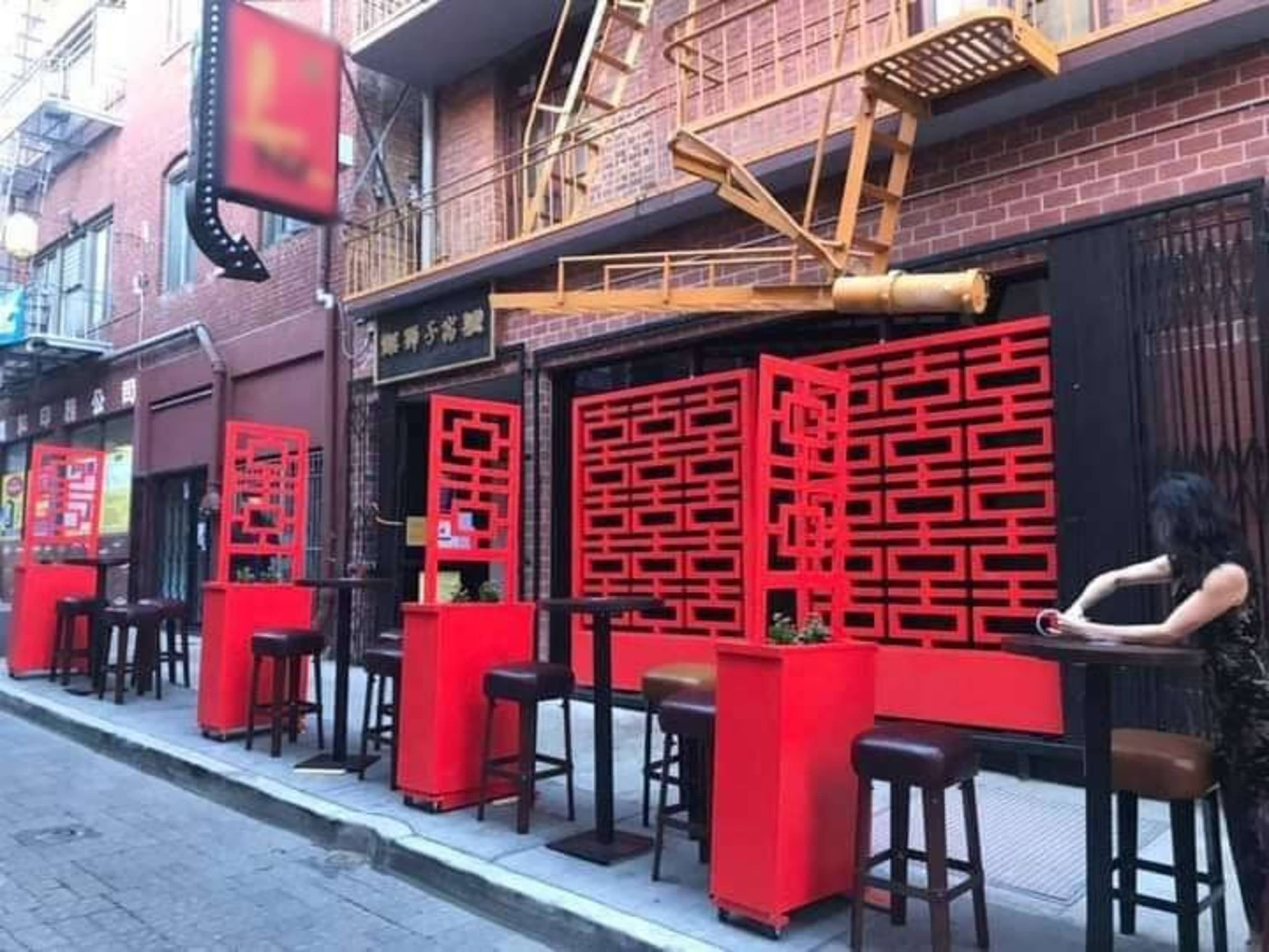 A narrow alley features outdoor seating with red decorative dividers and a woman seated at a high table.