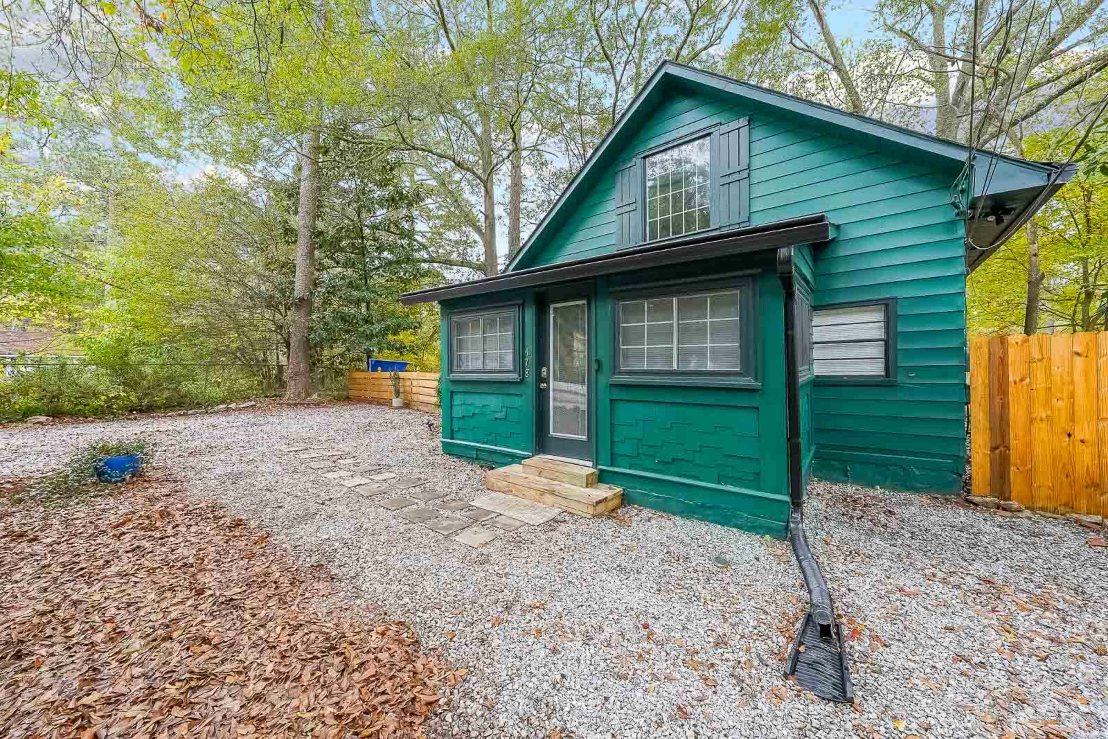 A green wooden cabin with a sloped roof is surrounded by gravel and trees in a secluded area.