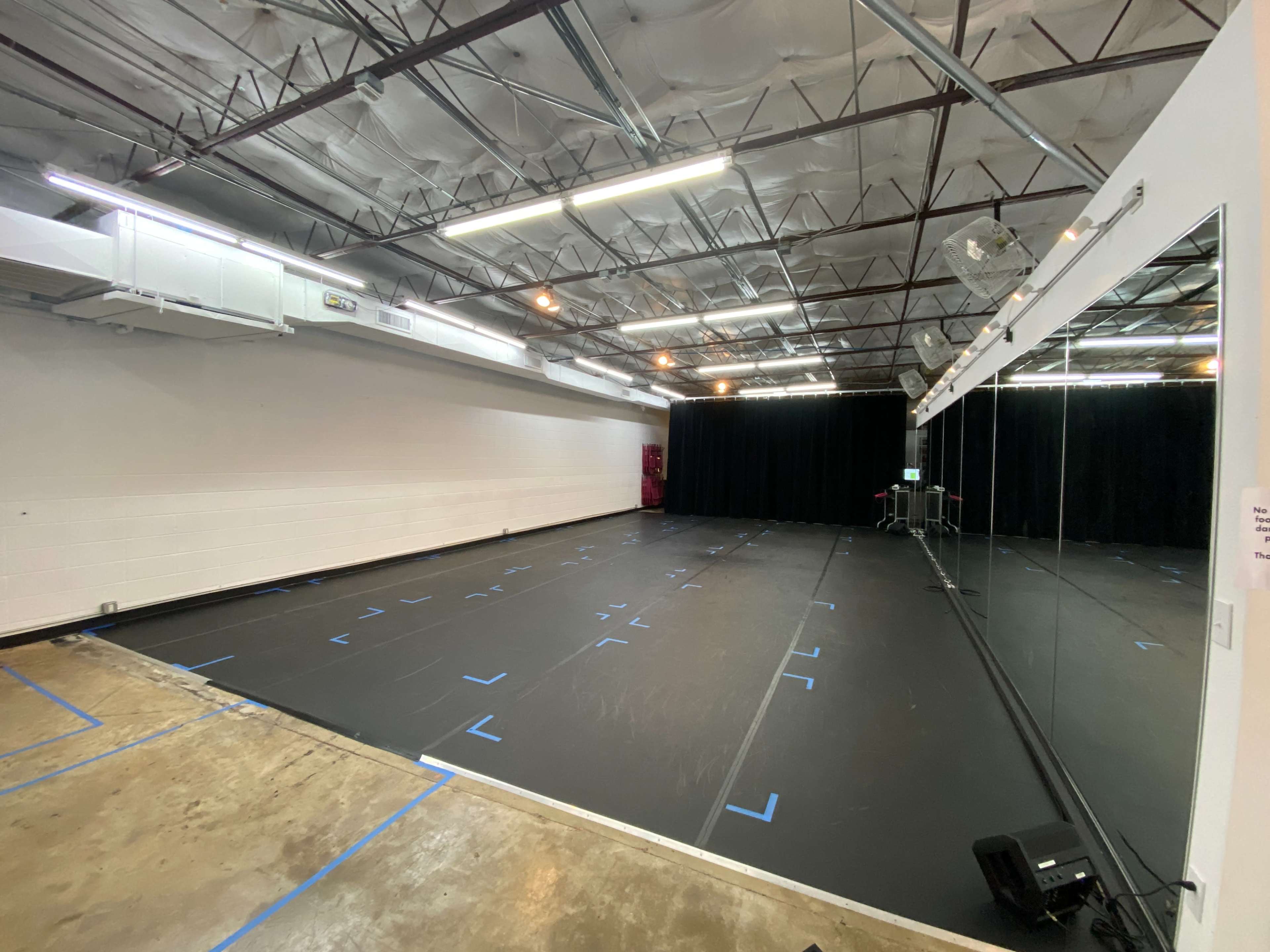 The image shows a spacious rehearsal studio with a black floor, mirrored wall, and a few pieces of fitness equipment in the corner.