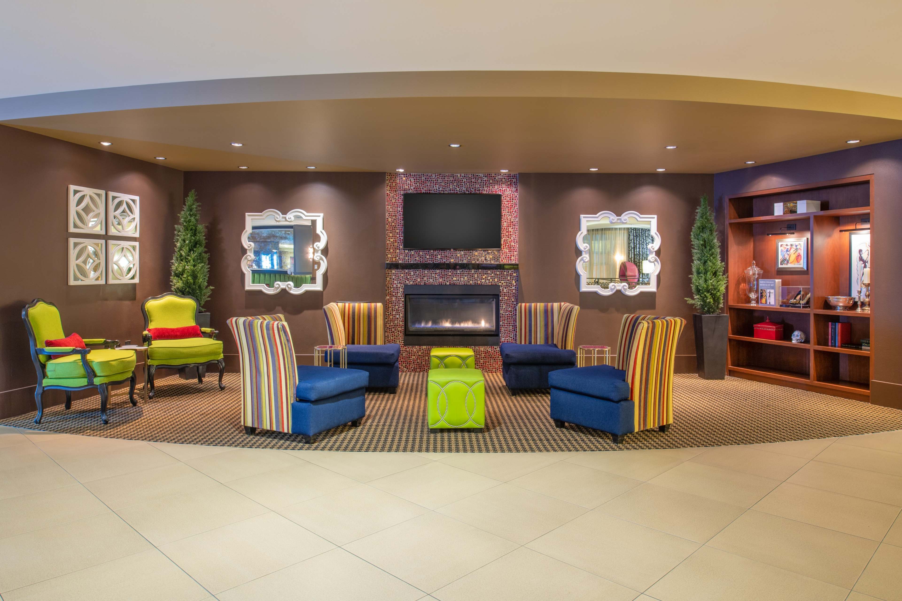 A lounge area with two striped chairs, two solid blue chairs, a central green table, a decorative fireplace, and a wall-mounted TV.