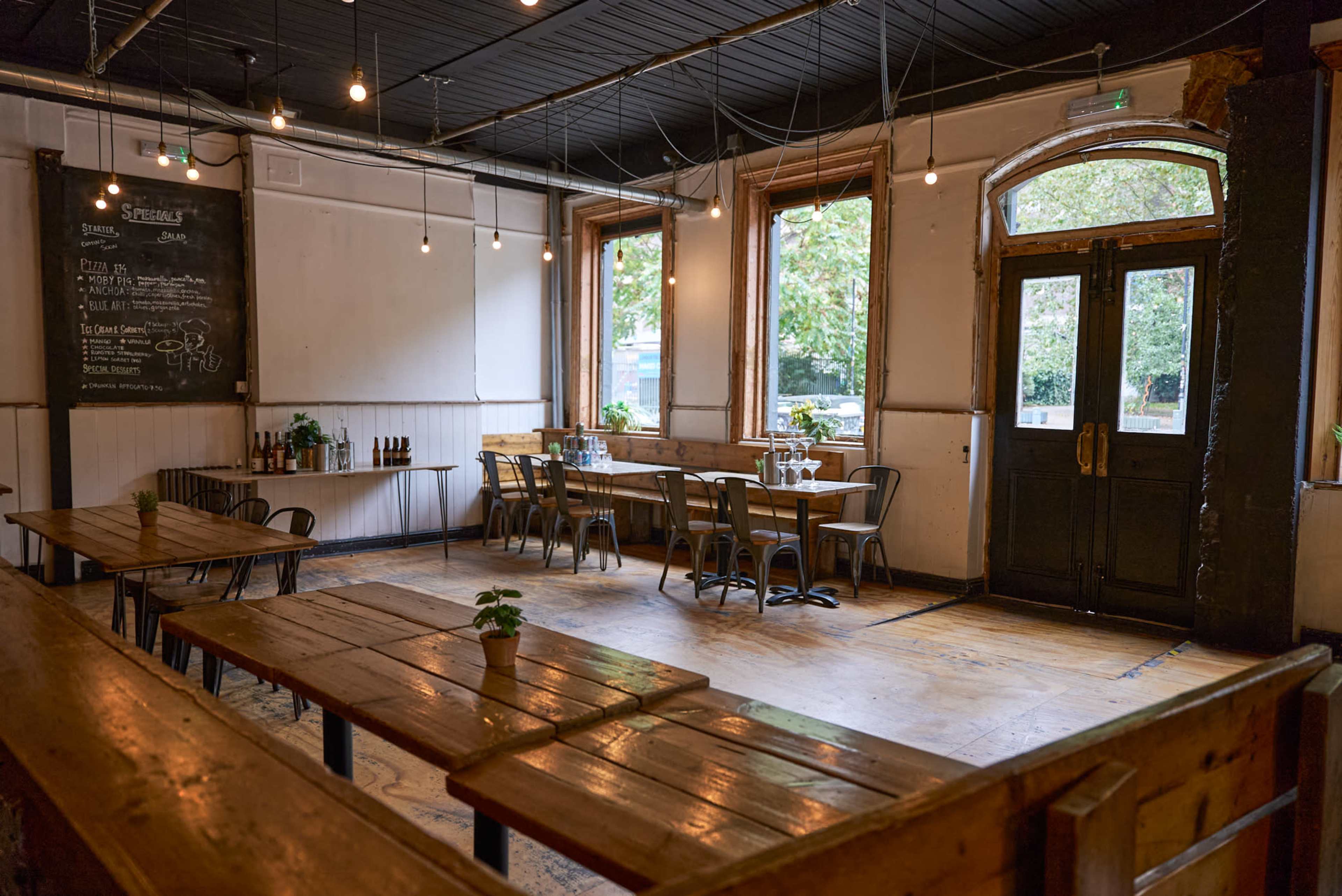 The image shows a rustic restaurant interior with wooden tables, chairs, and large windows allowing natural light to enter.