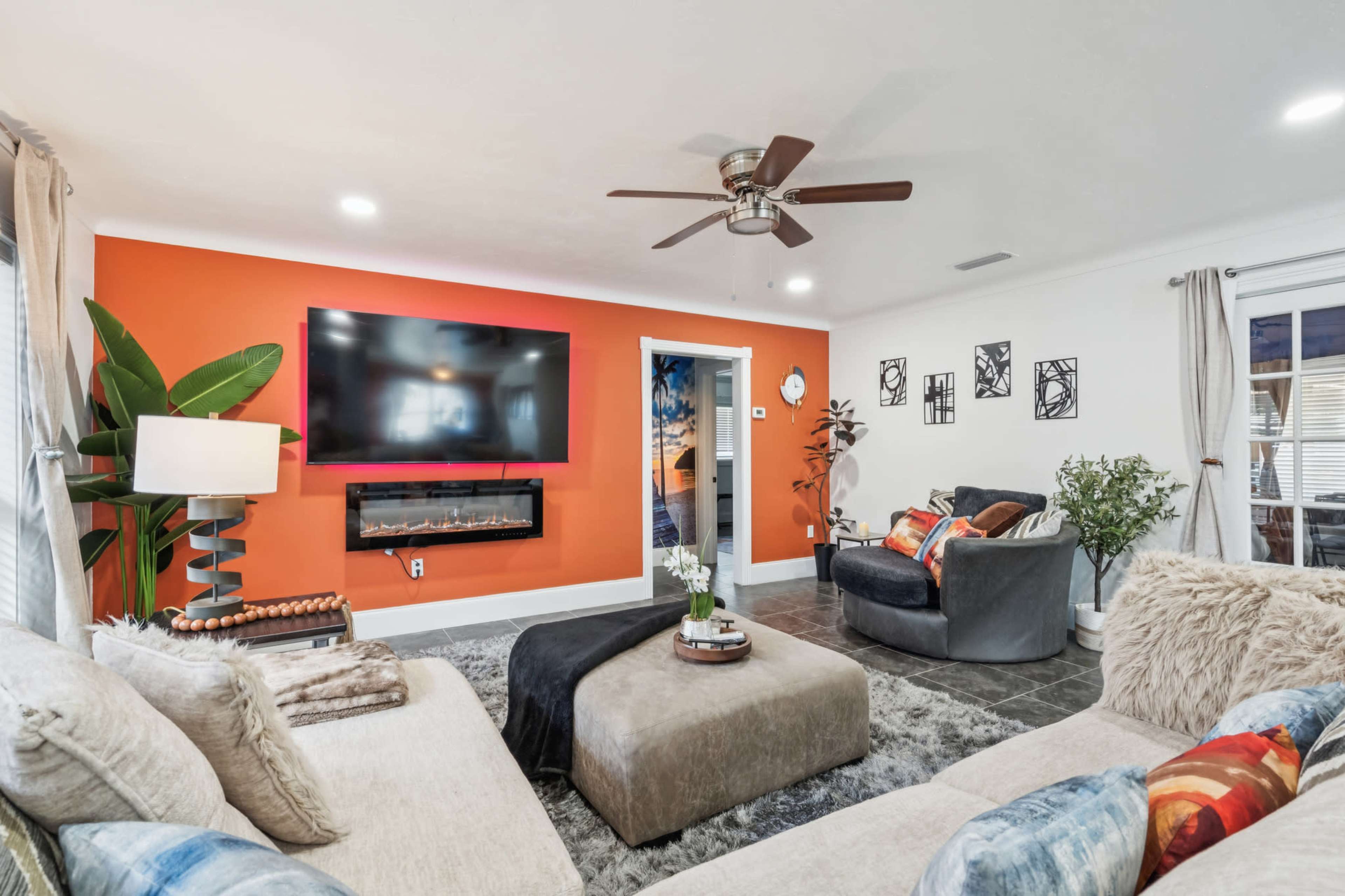 The living room features a gray sectional sofa, a wall-mounted TV above an electric fireplace, and an orange accent wall, with decorative plants and artwork.