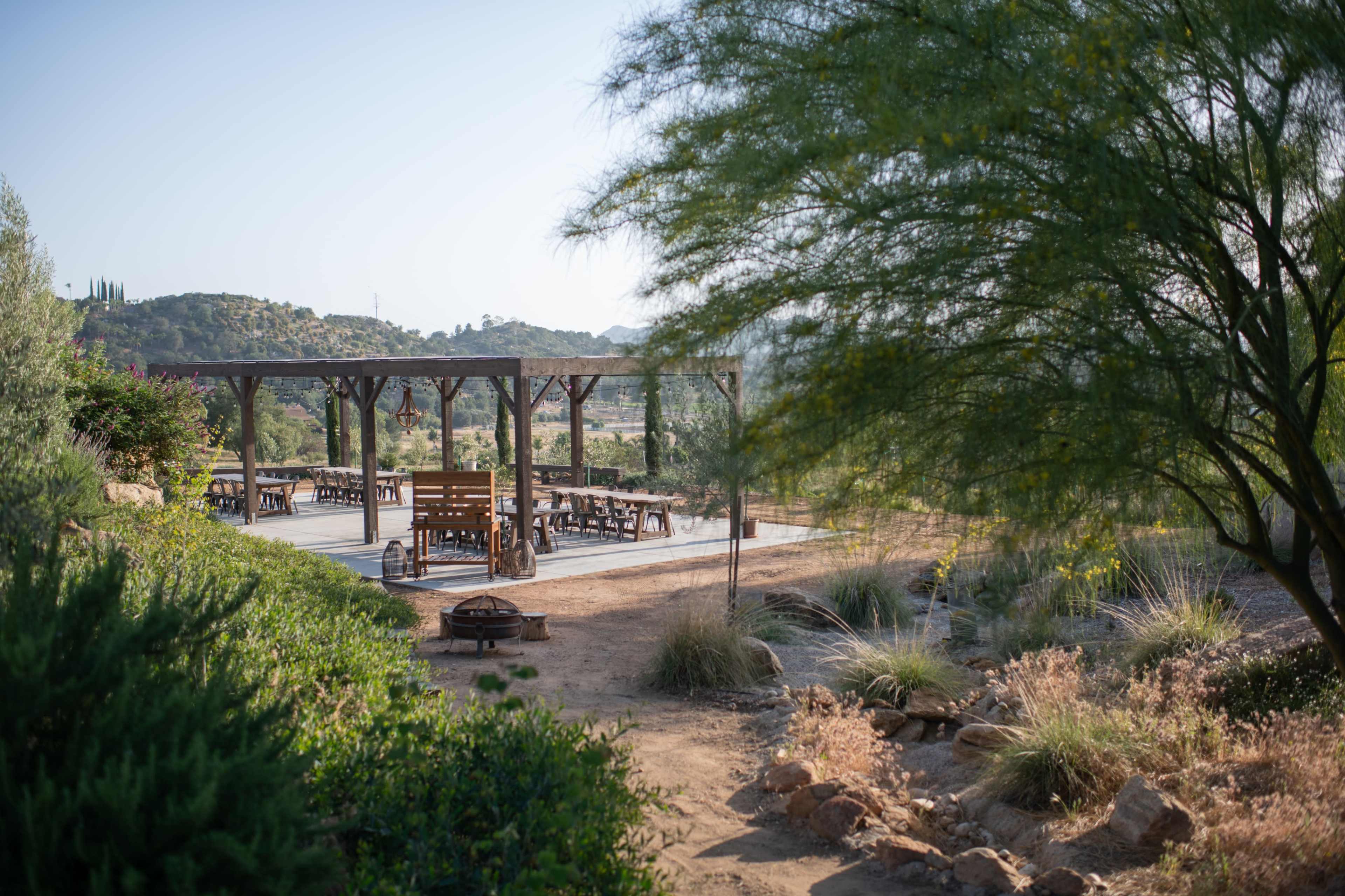 A wooden pavilion with seating surrounded by shrubs and mountains in the background.
