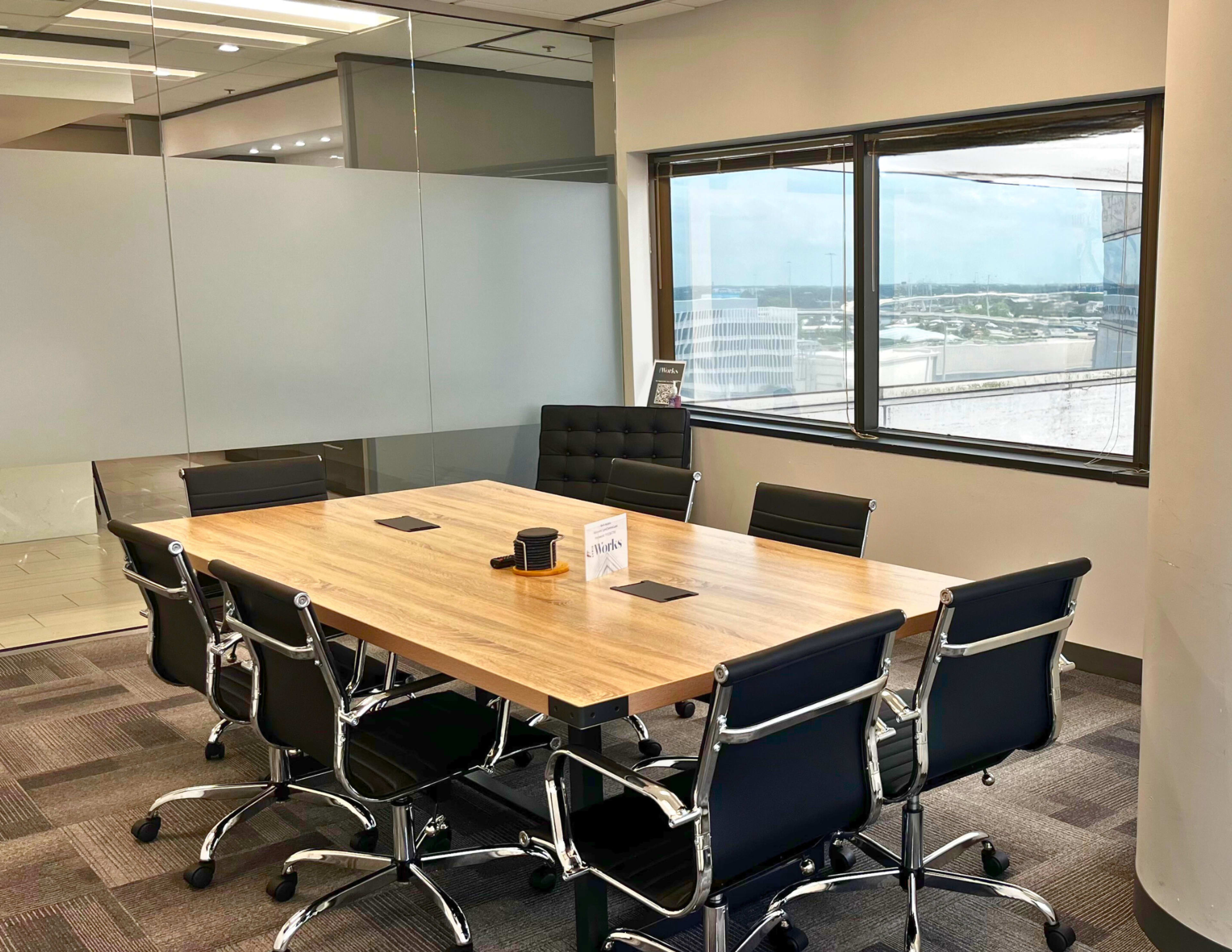 A conference room features a long wooden table surrounded by black office chairs, with large windows offering a view of the city.