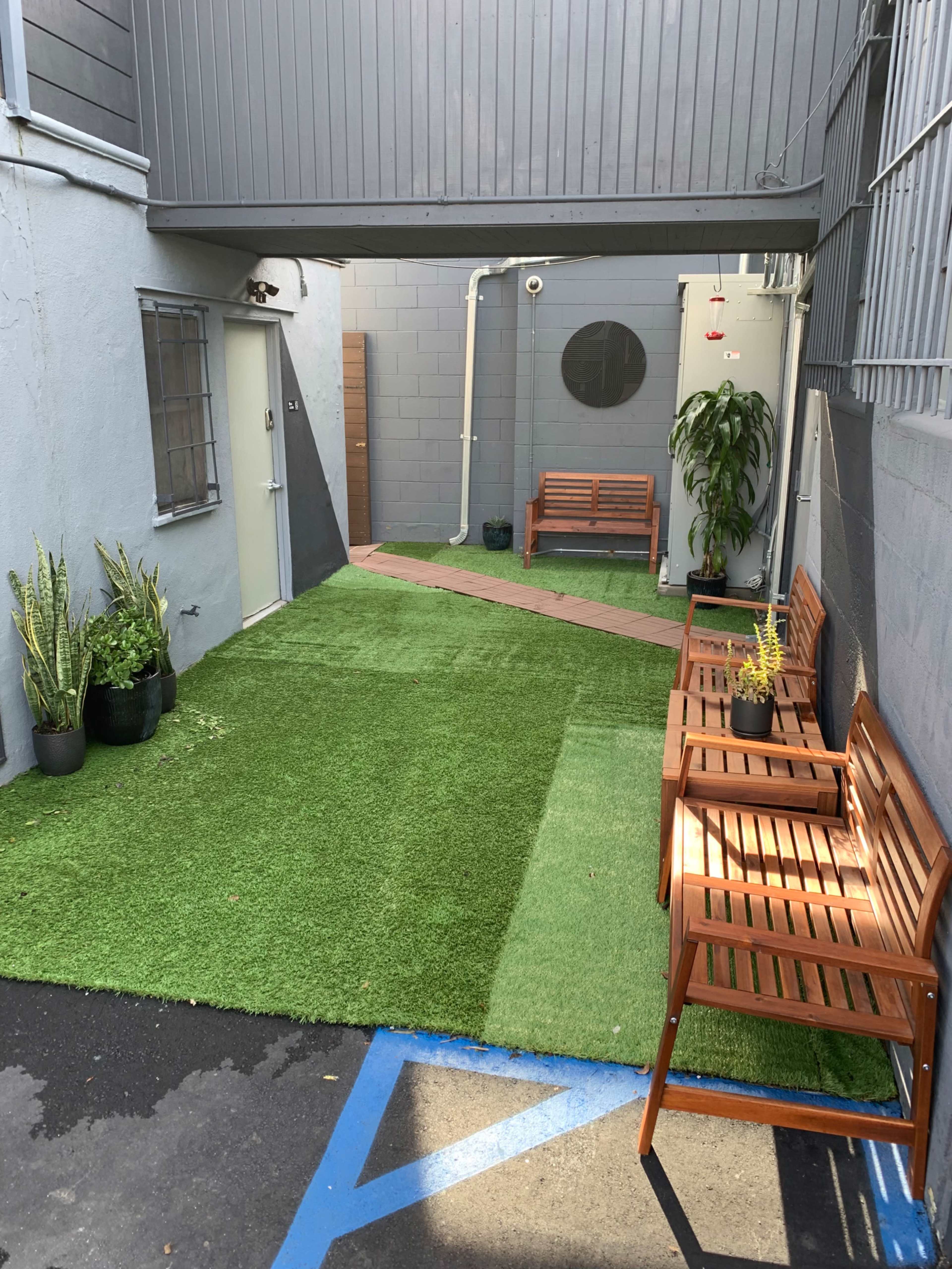 A narrow courtyard with artificial grass and wooden benches on either side, bordered by gray walls.