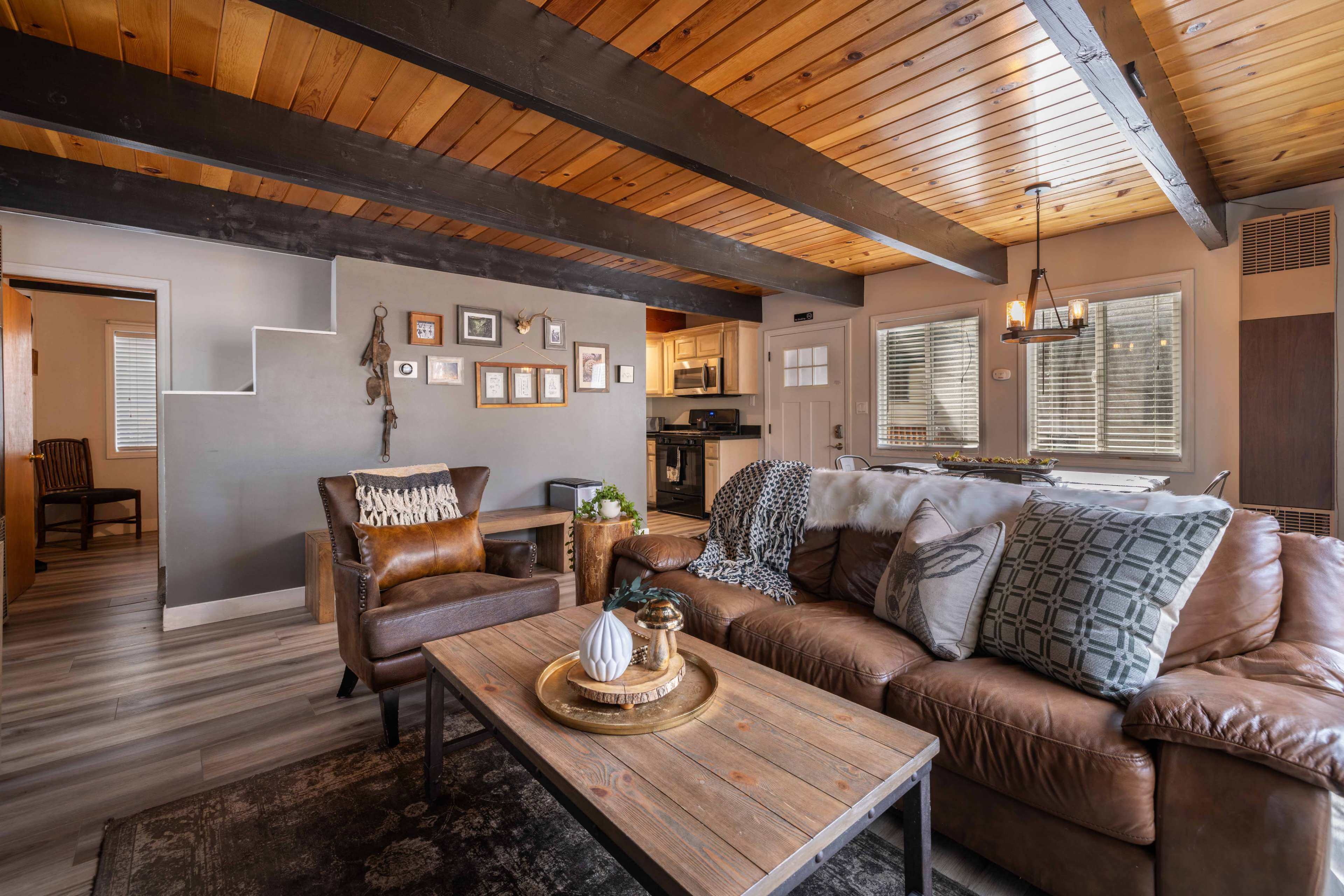 The image shows a cozy living room with a brown leather sofa, wooden ceiling beams, a coffee table with decorative items, and an open view into a kitchen area.