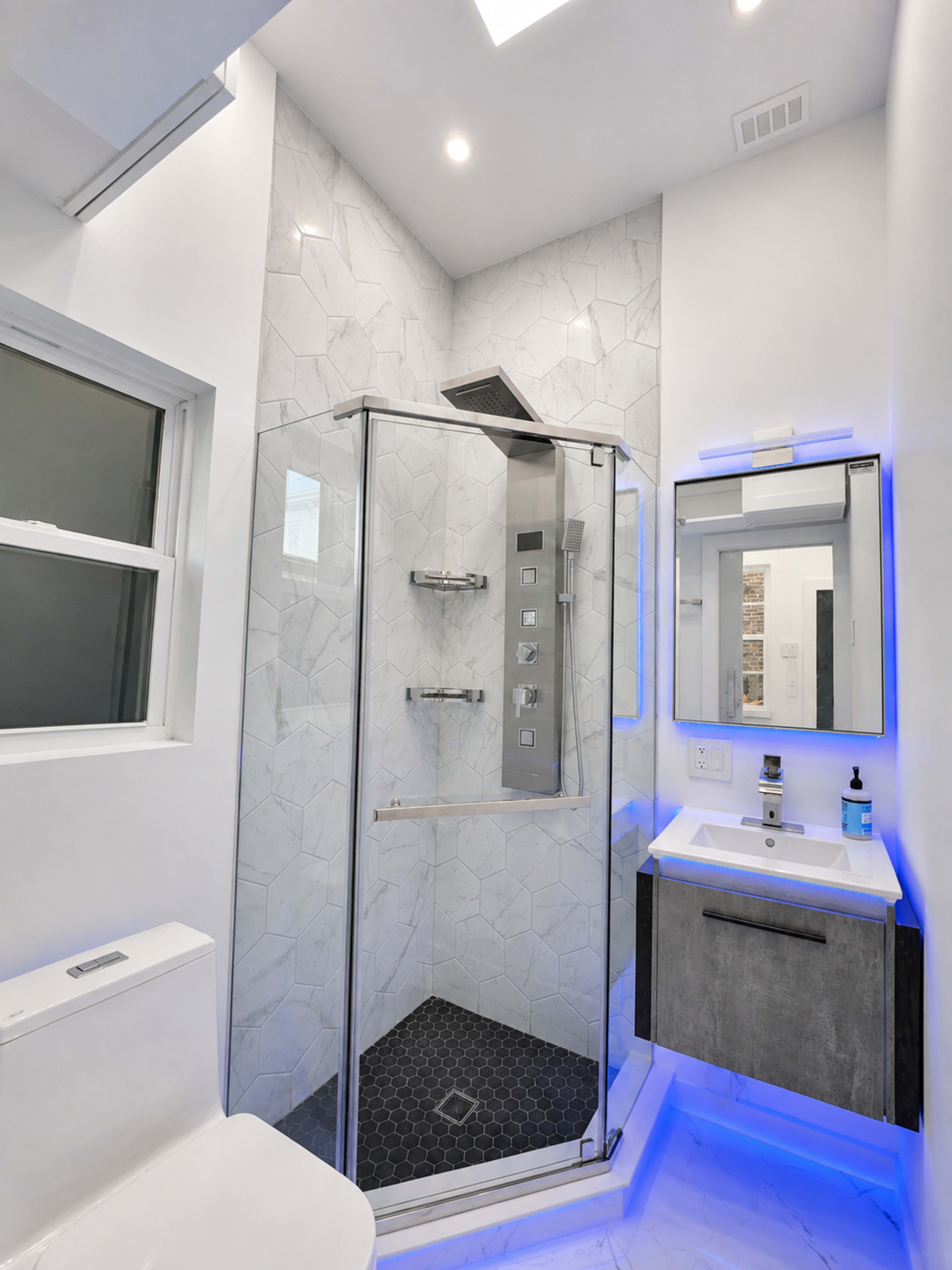 The image shows a modern bathroom featuring a glass shower enclosure with hexagonal tiles, a toilet, and a vanity with a mirror and backlit accents.