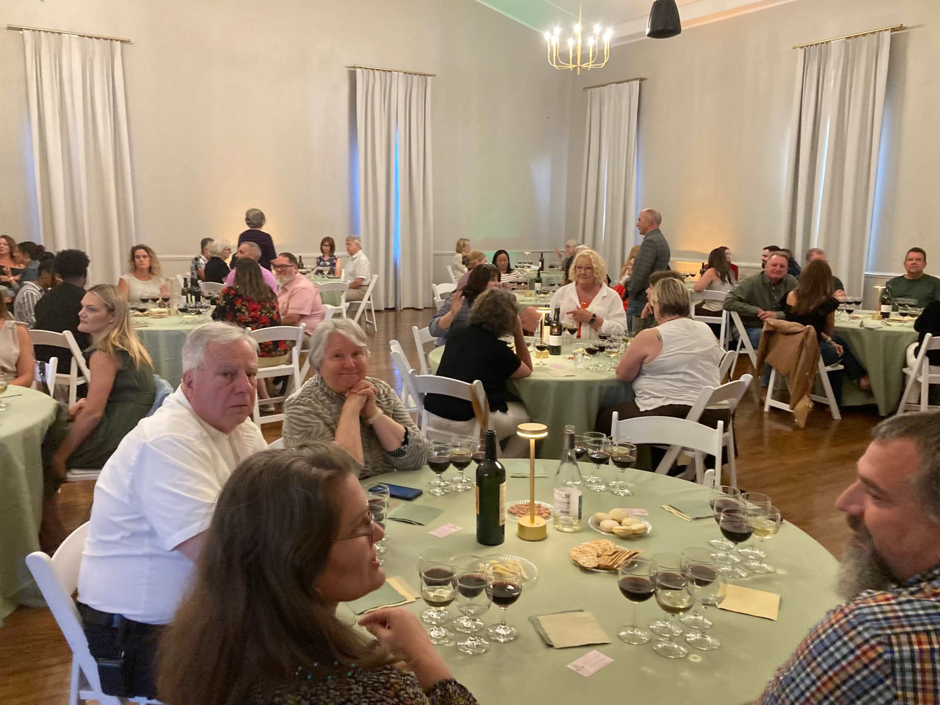 A large group of people is seated at round tables in a banquet hall, enjoying drinks and conversation.