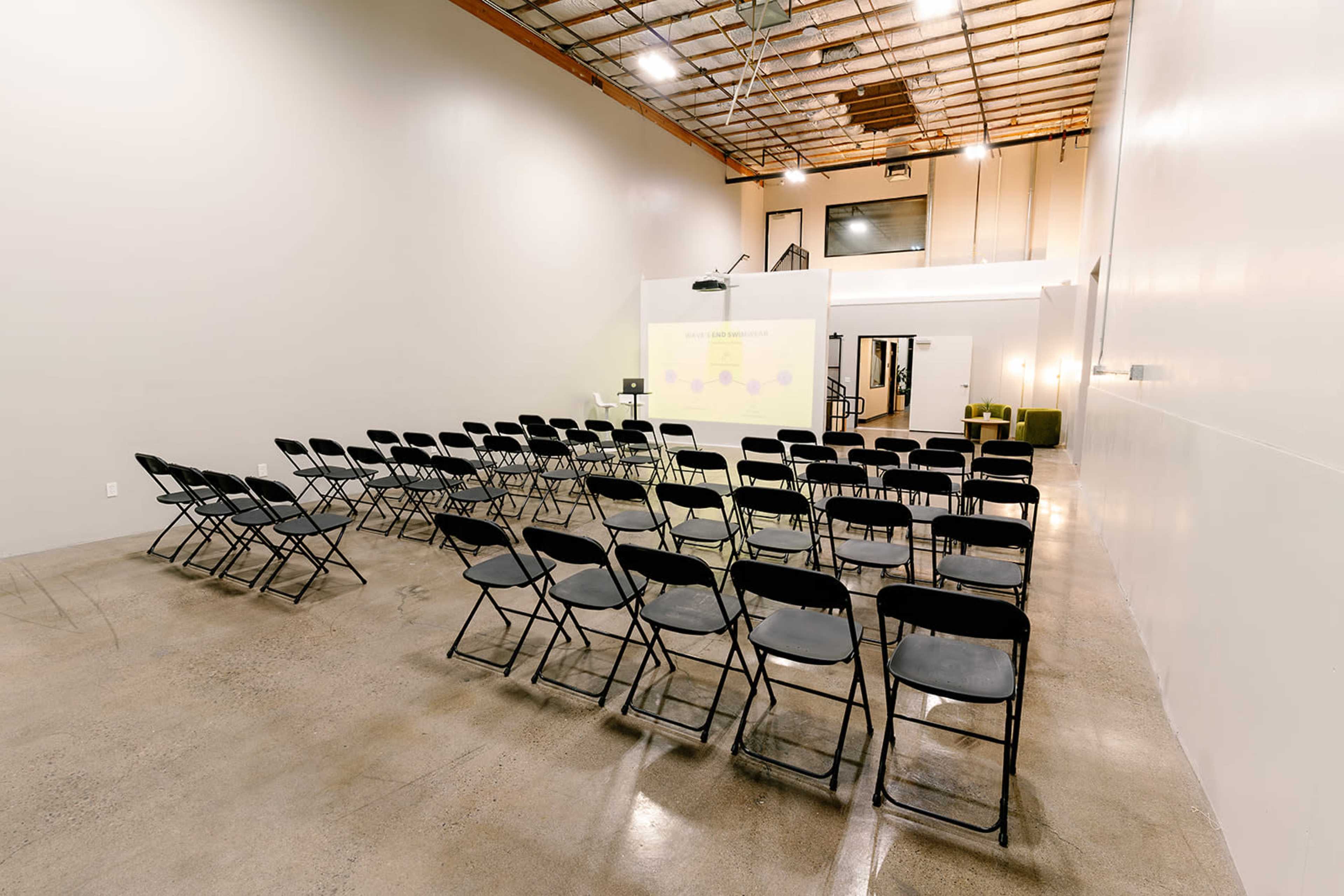 A spacious room with a polished concrete floor is set up with rows of black folding chairs facing a screen at the front.