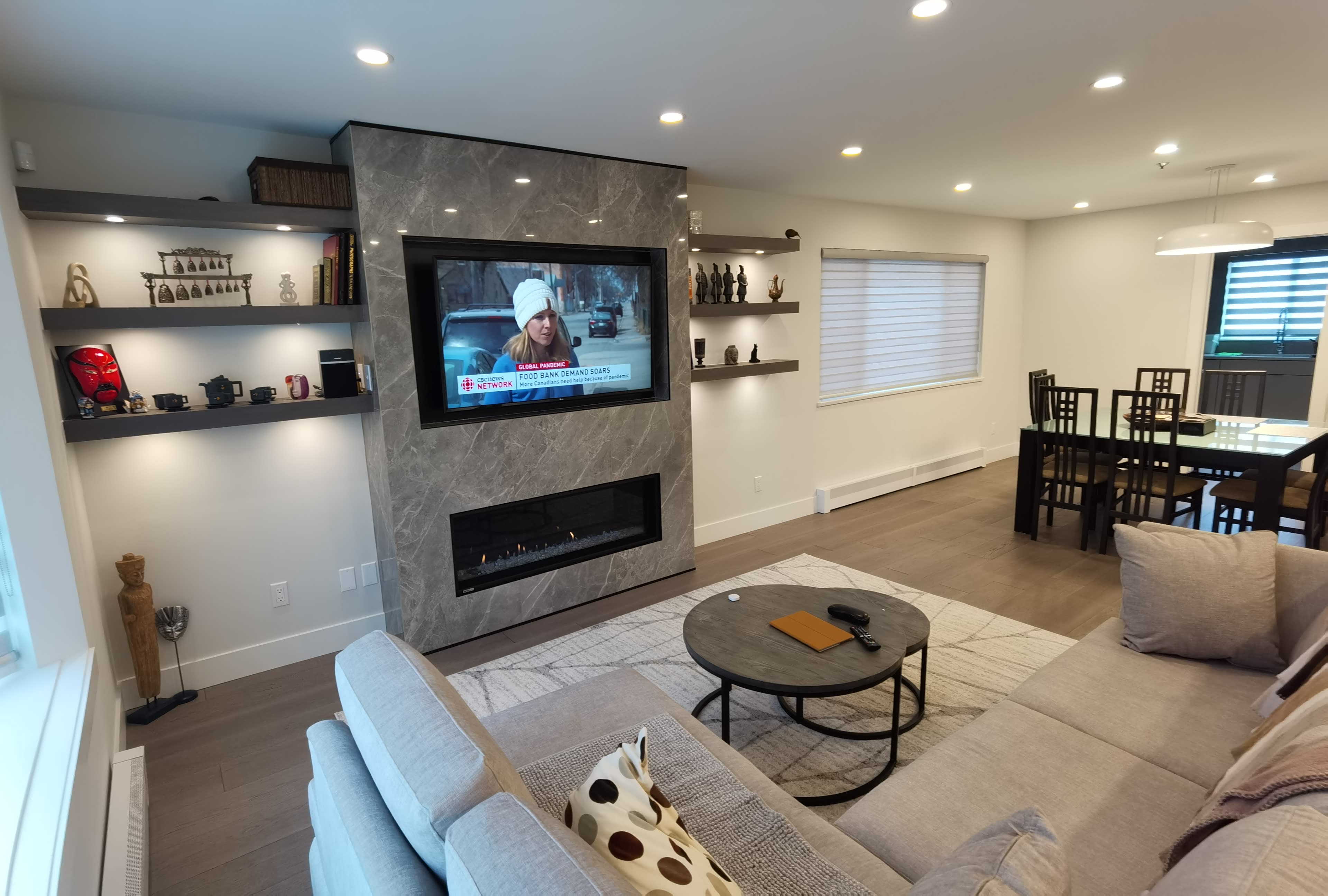 A modern living room with a gray stone fireplace, a large television displaying a news broadcast, and a dining area in the background.