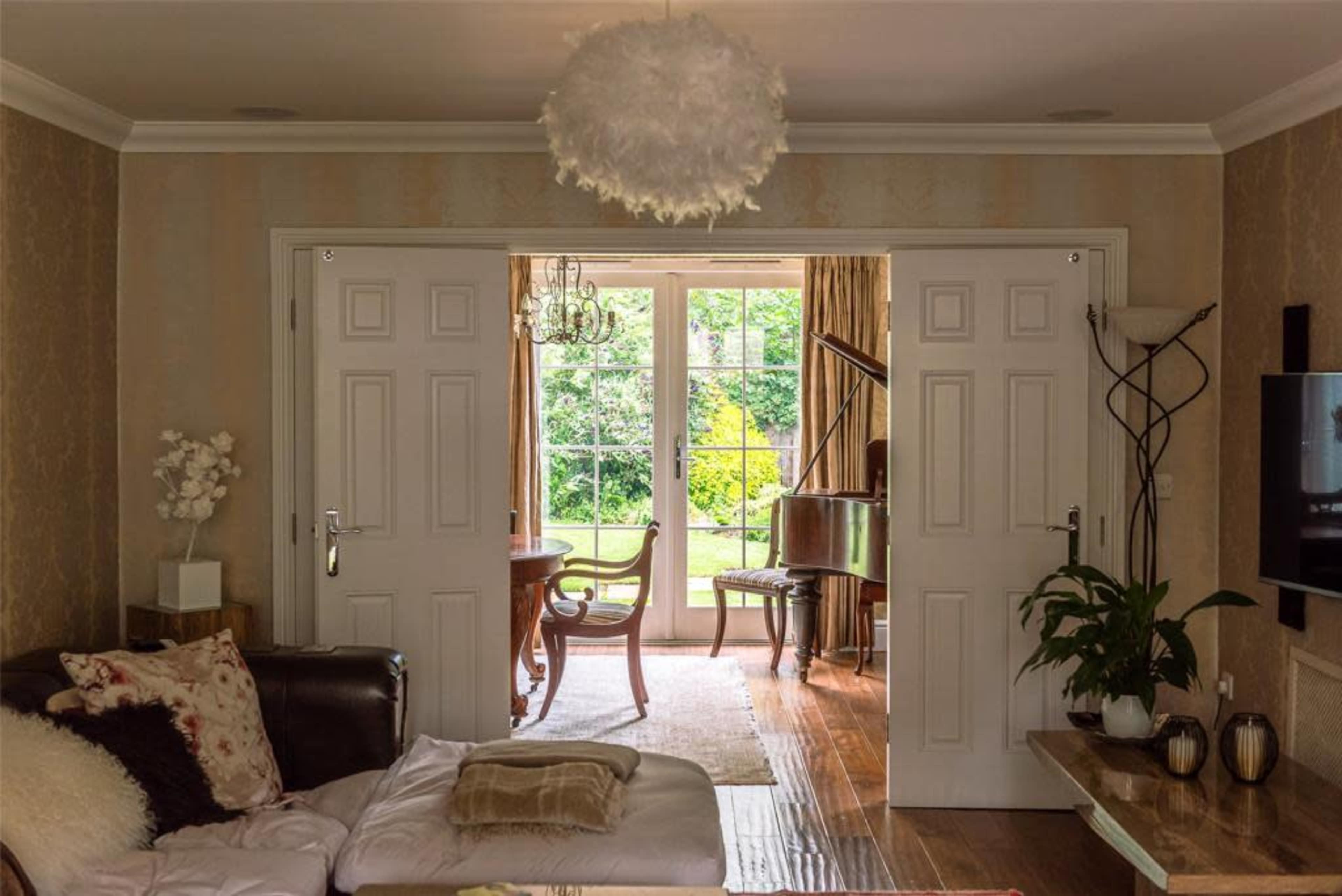A living room with a brown couch and decorative pillows leads to a sunlit dining area featuring a piano and large windows overlooking a garden.