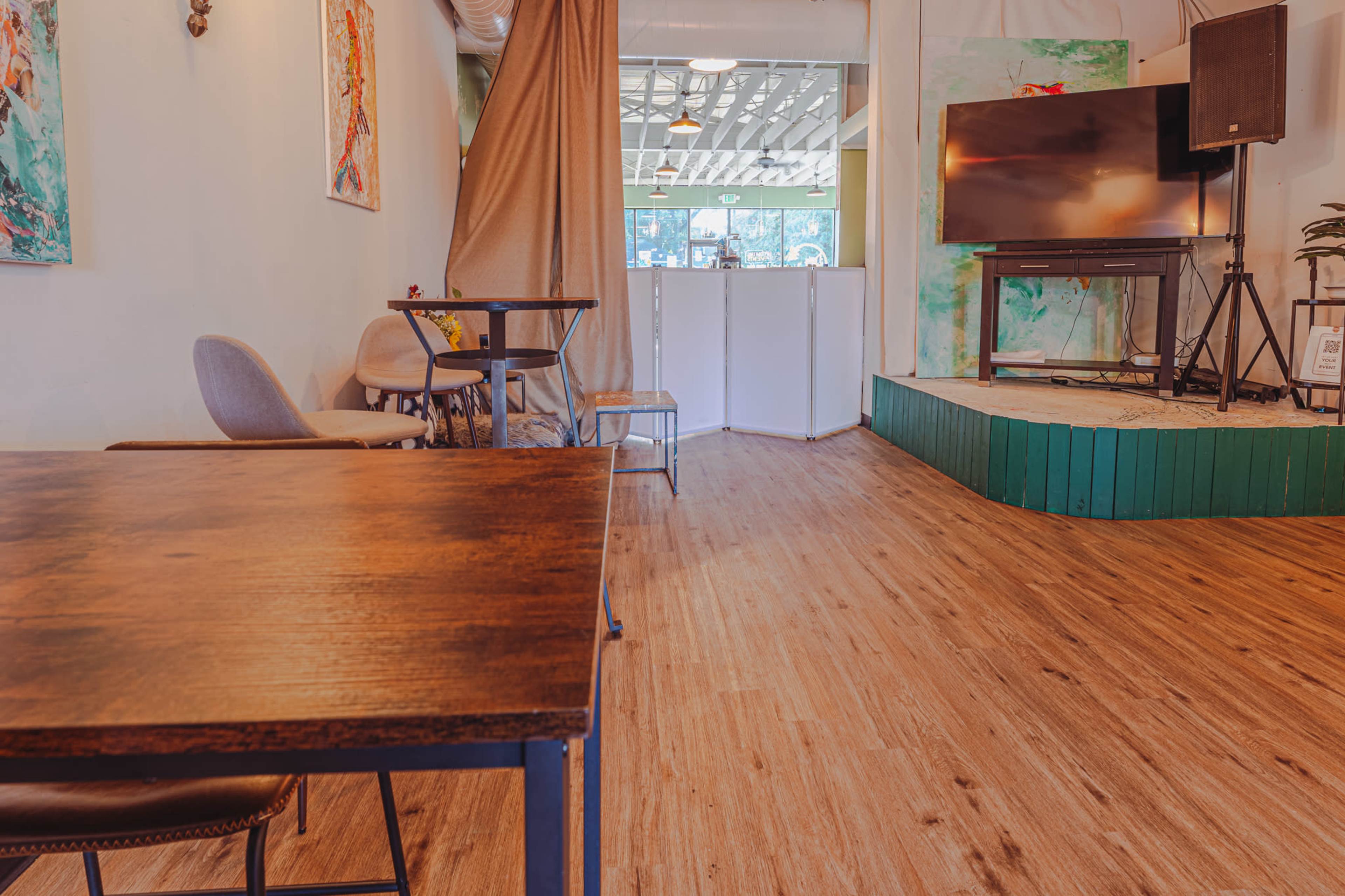 The image shows a cafe interior featuring wooden flooring, a small table and chairs, and a raised green platform with a television in the background.