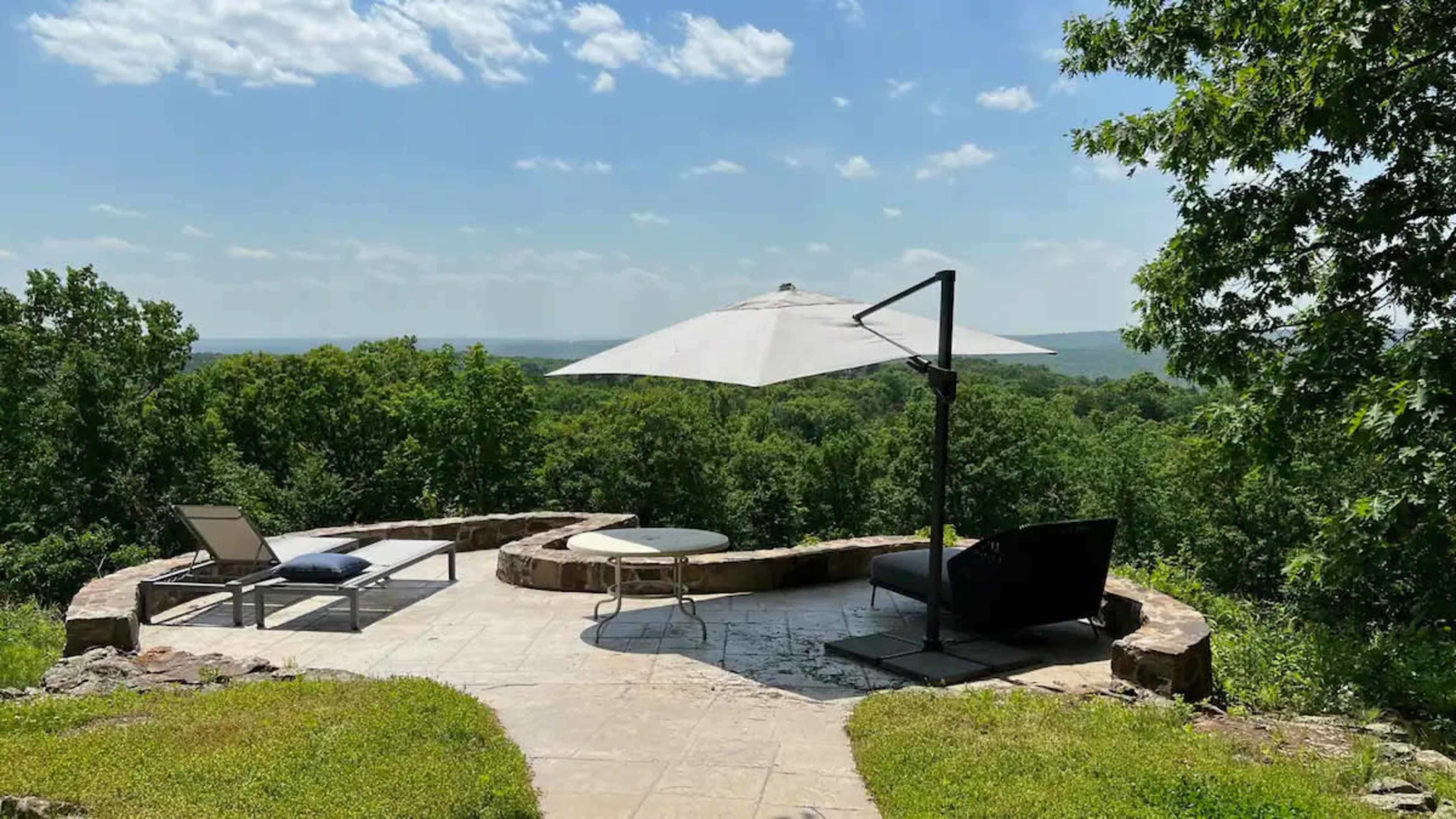 The image shows a patio with a seating area, an umbrella, and a view of dense greenery and a distant landscape under a blue sky.