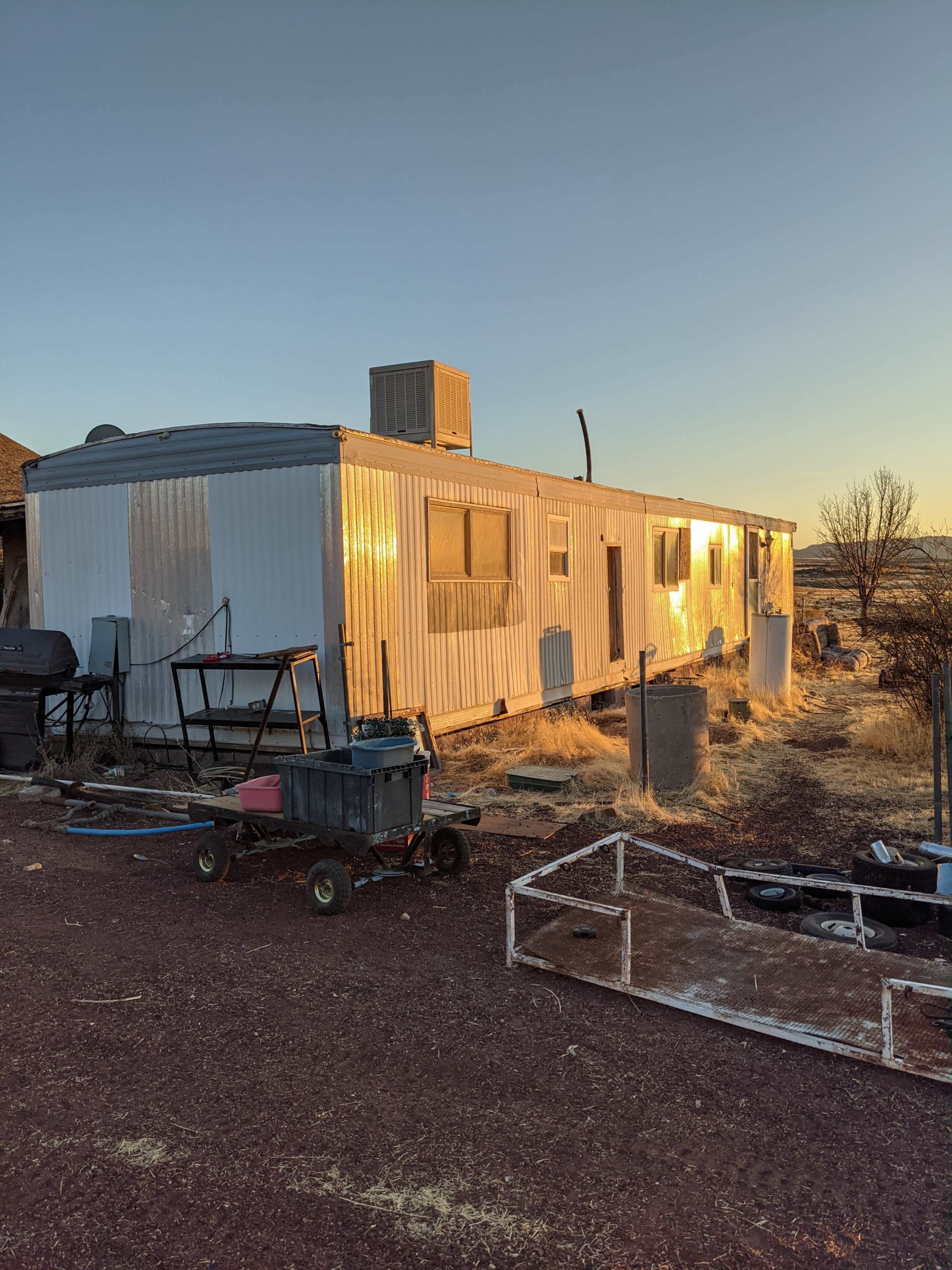 A metal-sided mobile home sits on a gravel lot, with various tools and equipment nearby.