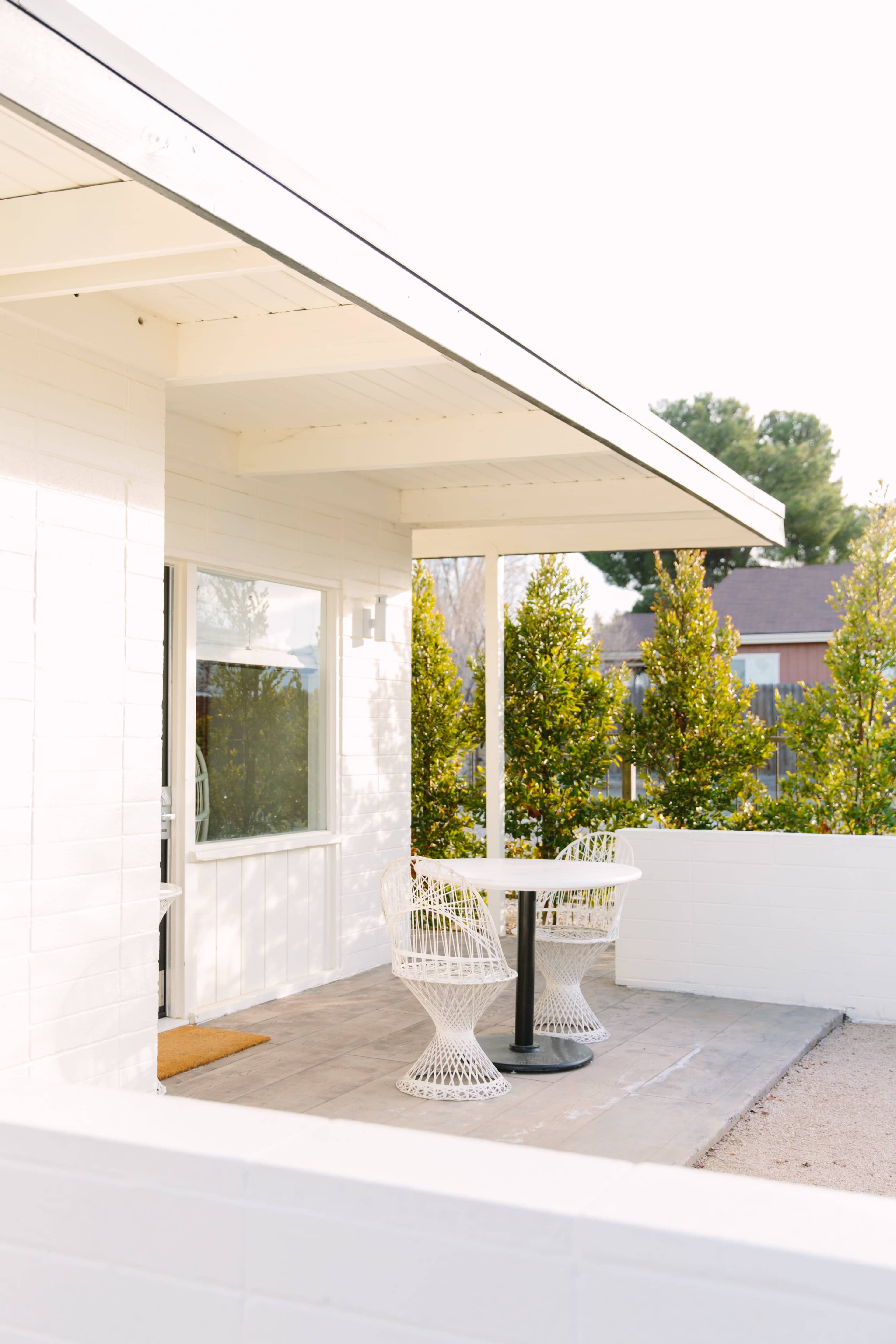 A white patio features a round table surrounded by two woven chairs under a covered area, bordered by greenery.