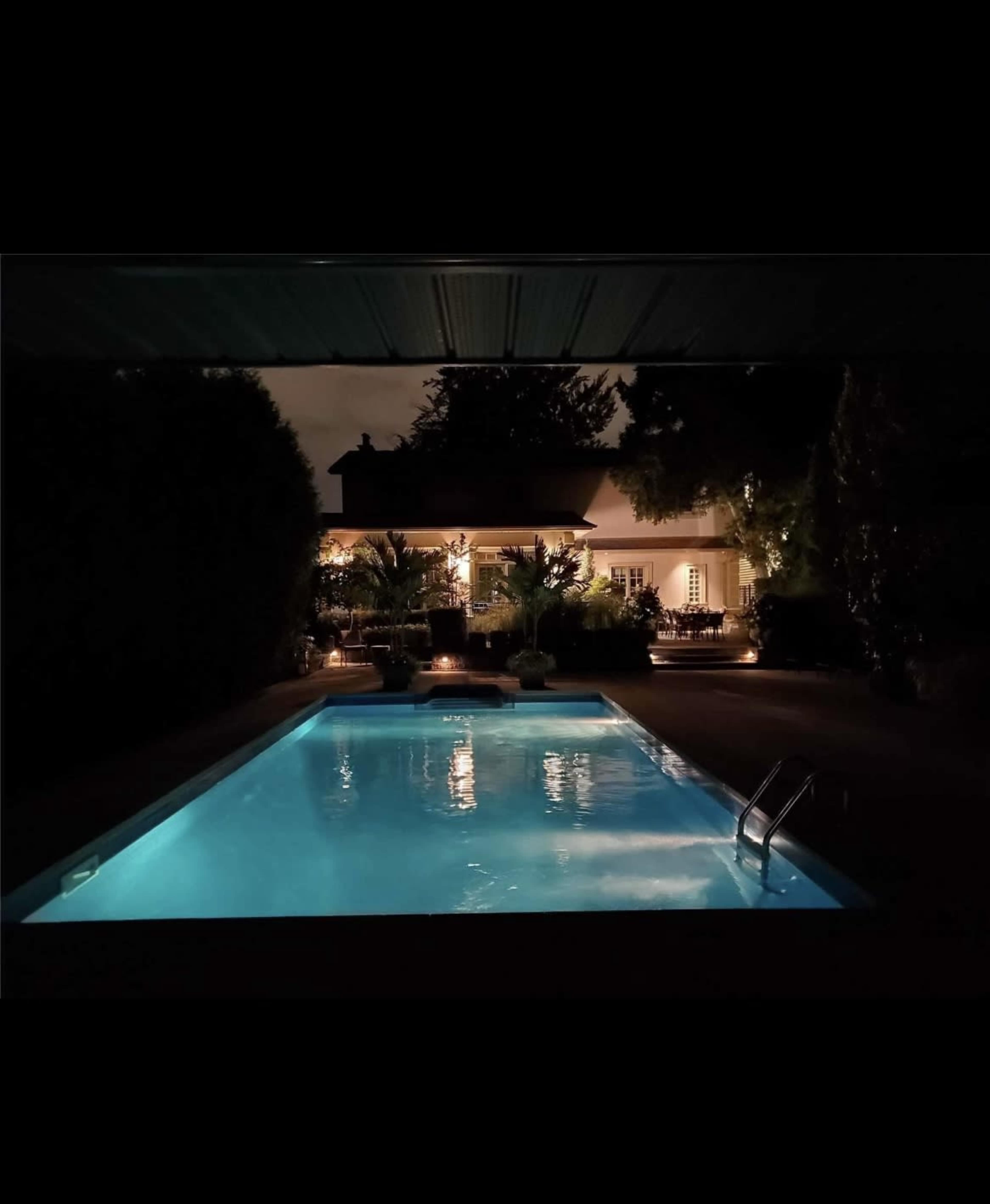 A serene swimming pool is illuminated at night, surrounded by lush greenery and a well-lit house in the background.