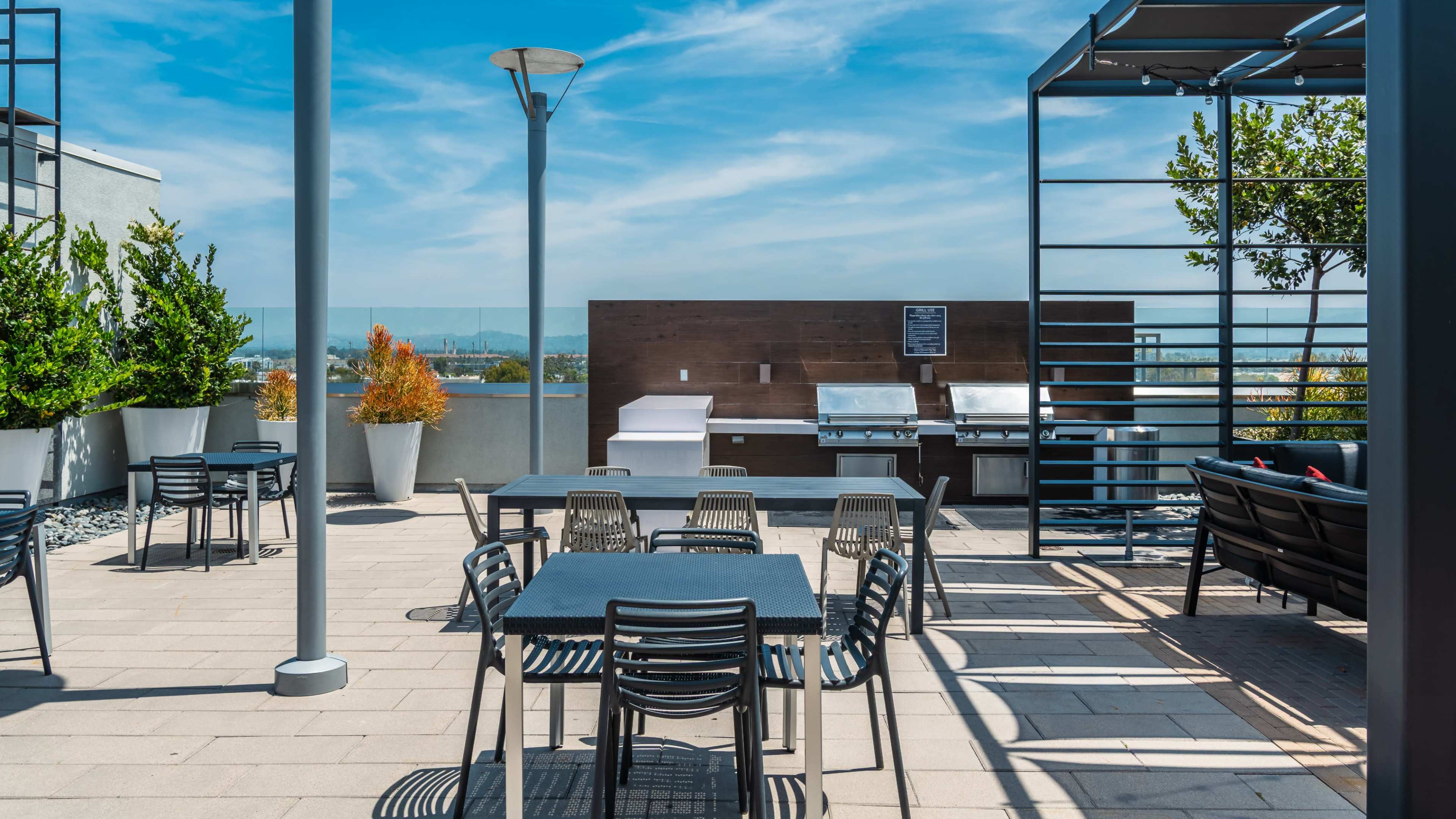 The image shows a rooftop area with several tables and chairs, a grilling station, plants in large pots, and a clear blue sky.