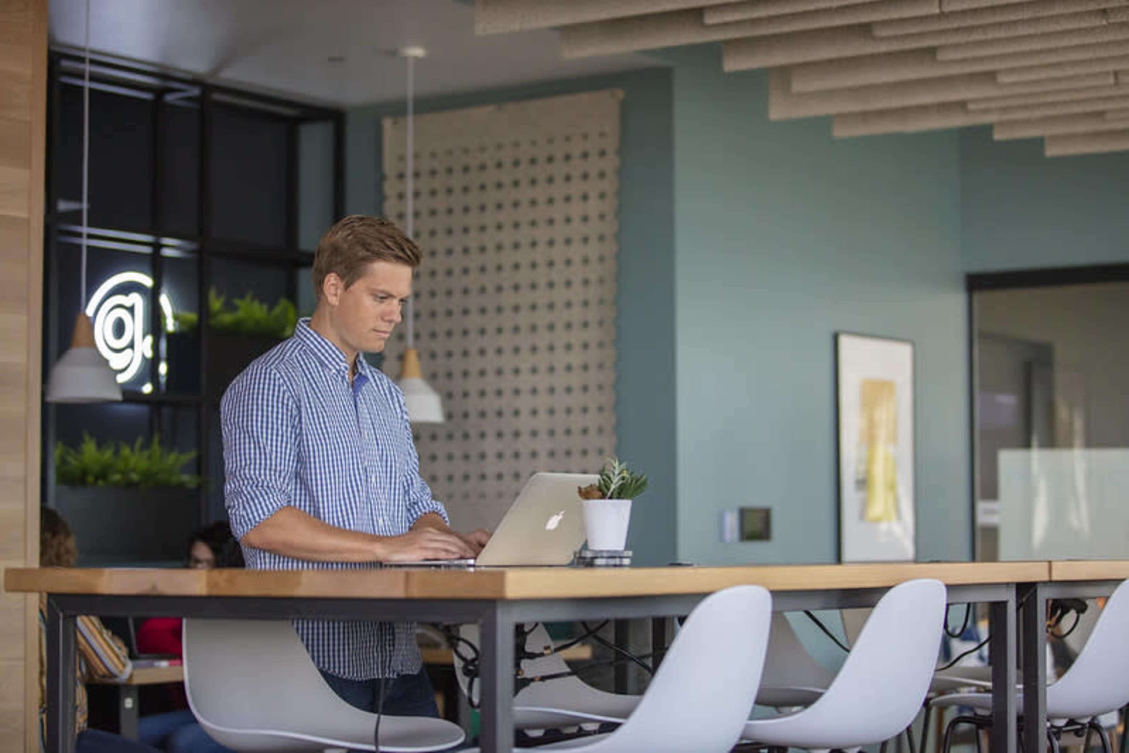 A man in a checked shirt stands at a table working on a laptop in a modern, well-lit workspace.