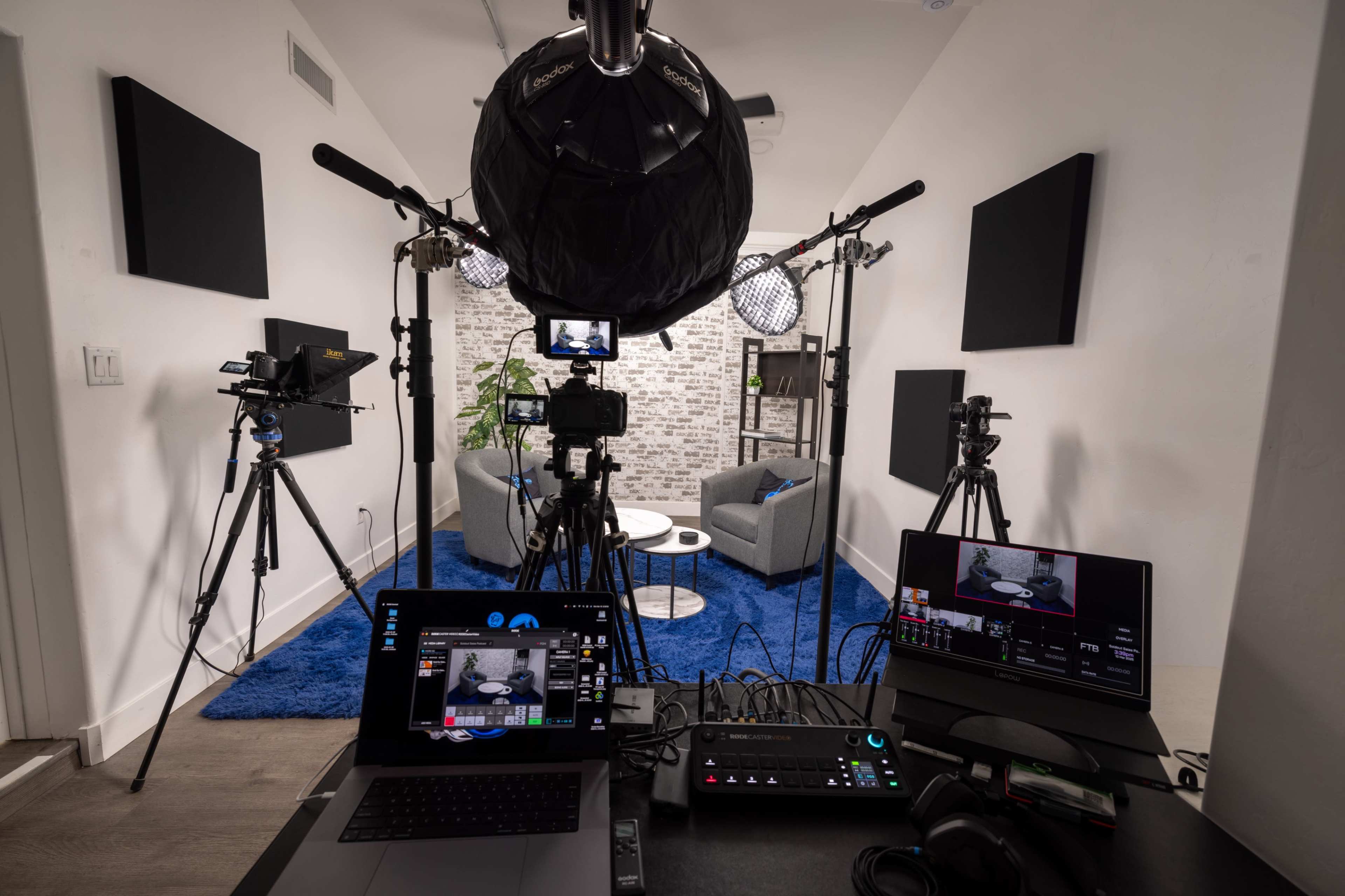The image shows a modern video production setup in a studio with cameras, lighting equipment, and a computer displaying a live feed, surrounded by a seating area.