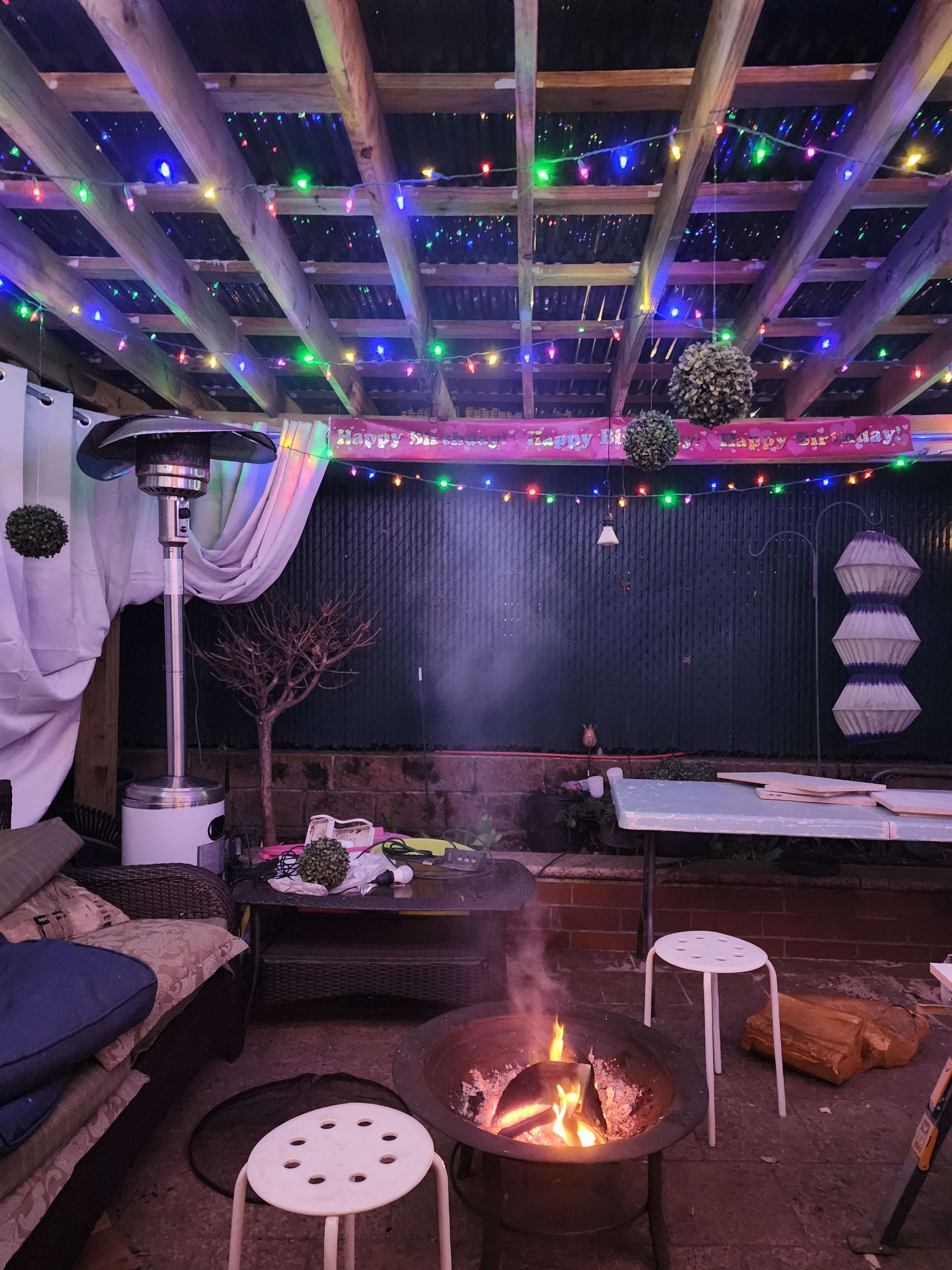The scene features a cozy outdoor seating area with colorful lights strung overhead, a fire pit in the center, and various items scattered on the ground.