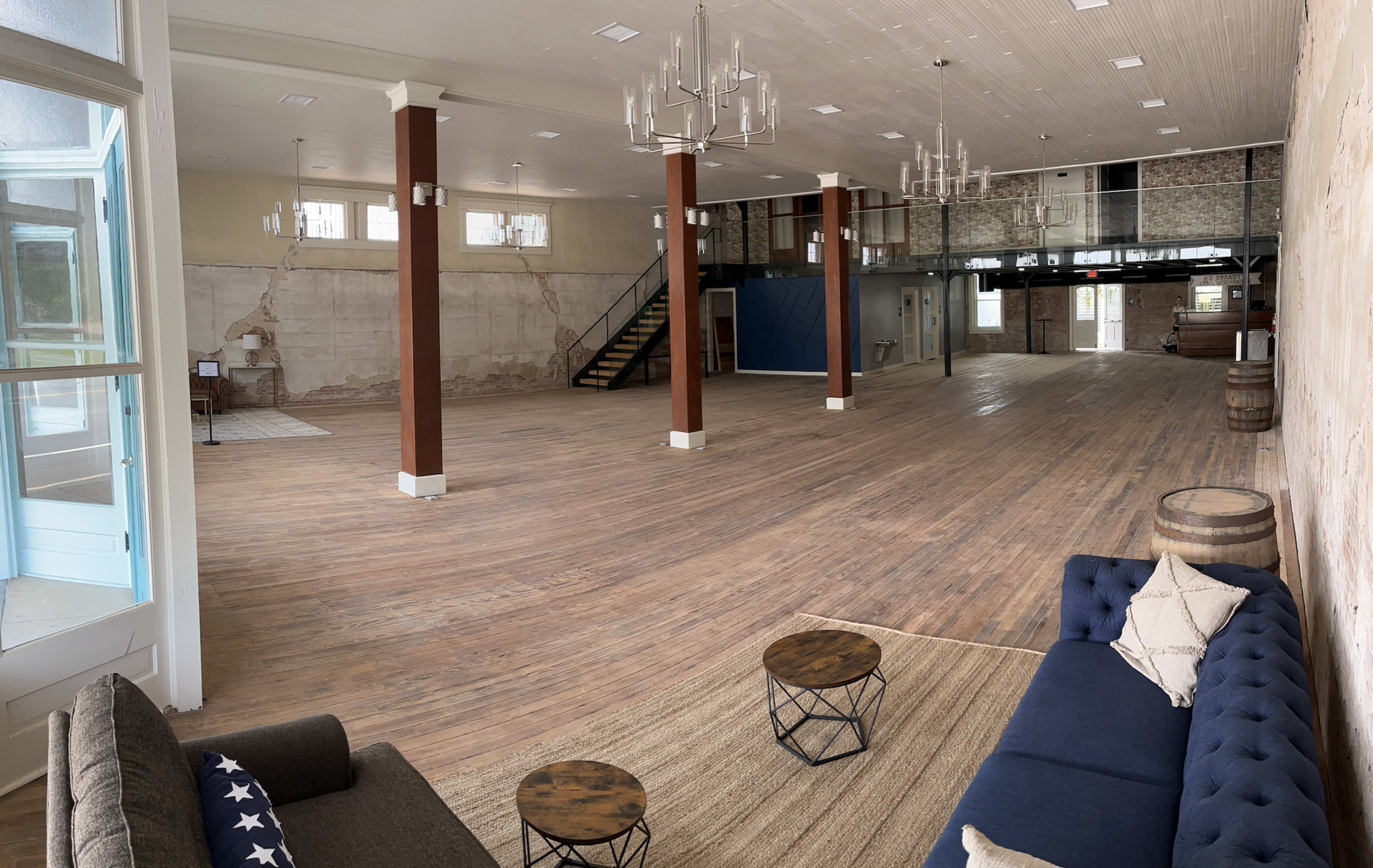 The image shows a spacious, empty interior with wooden floors, exposed walls, and a seating area featuring a dark blue sofa and small tables.