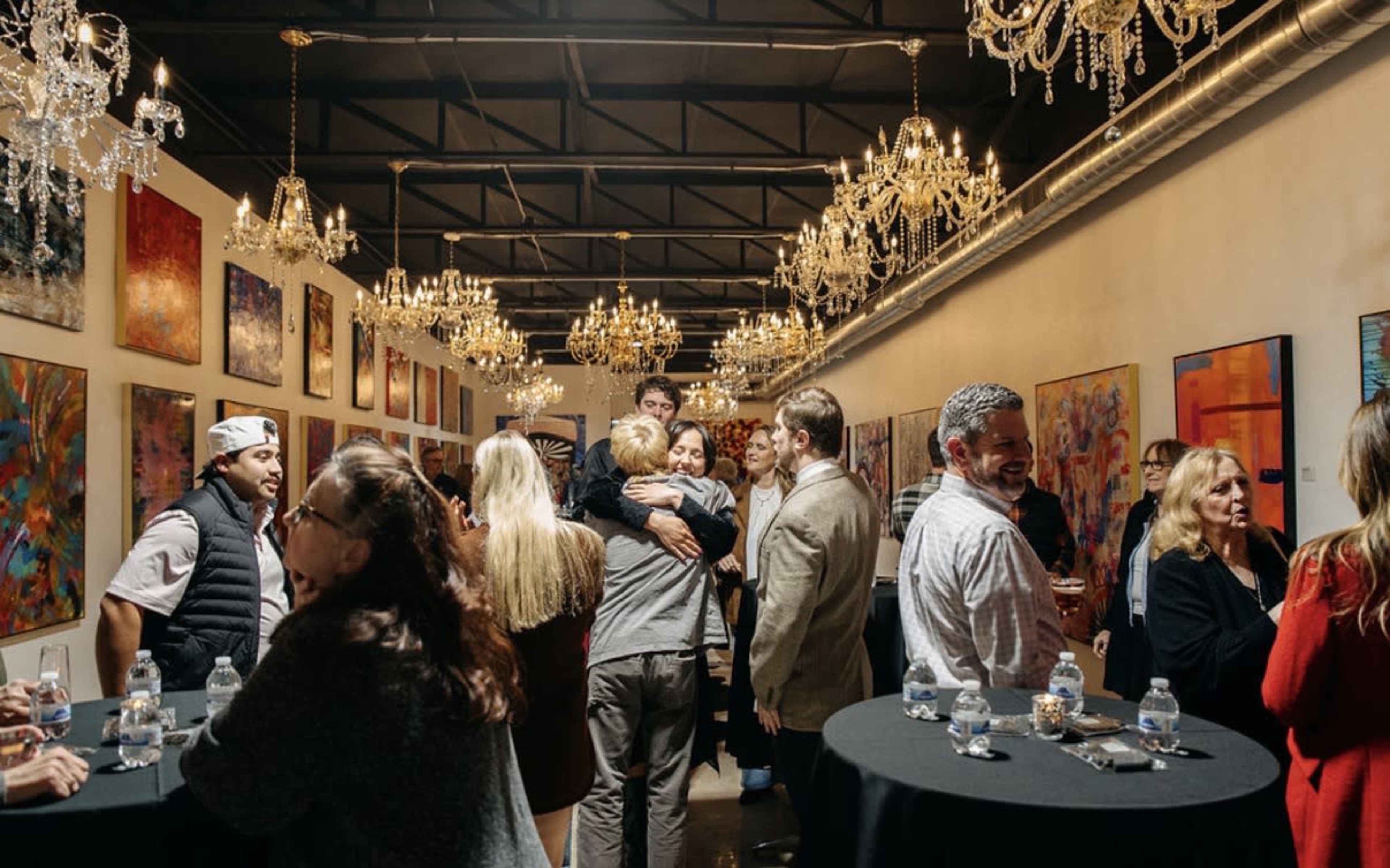 A group of people mingles in an art gallery adorned with chandeliers and colorful paintings on the walls.