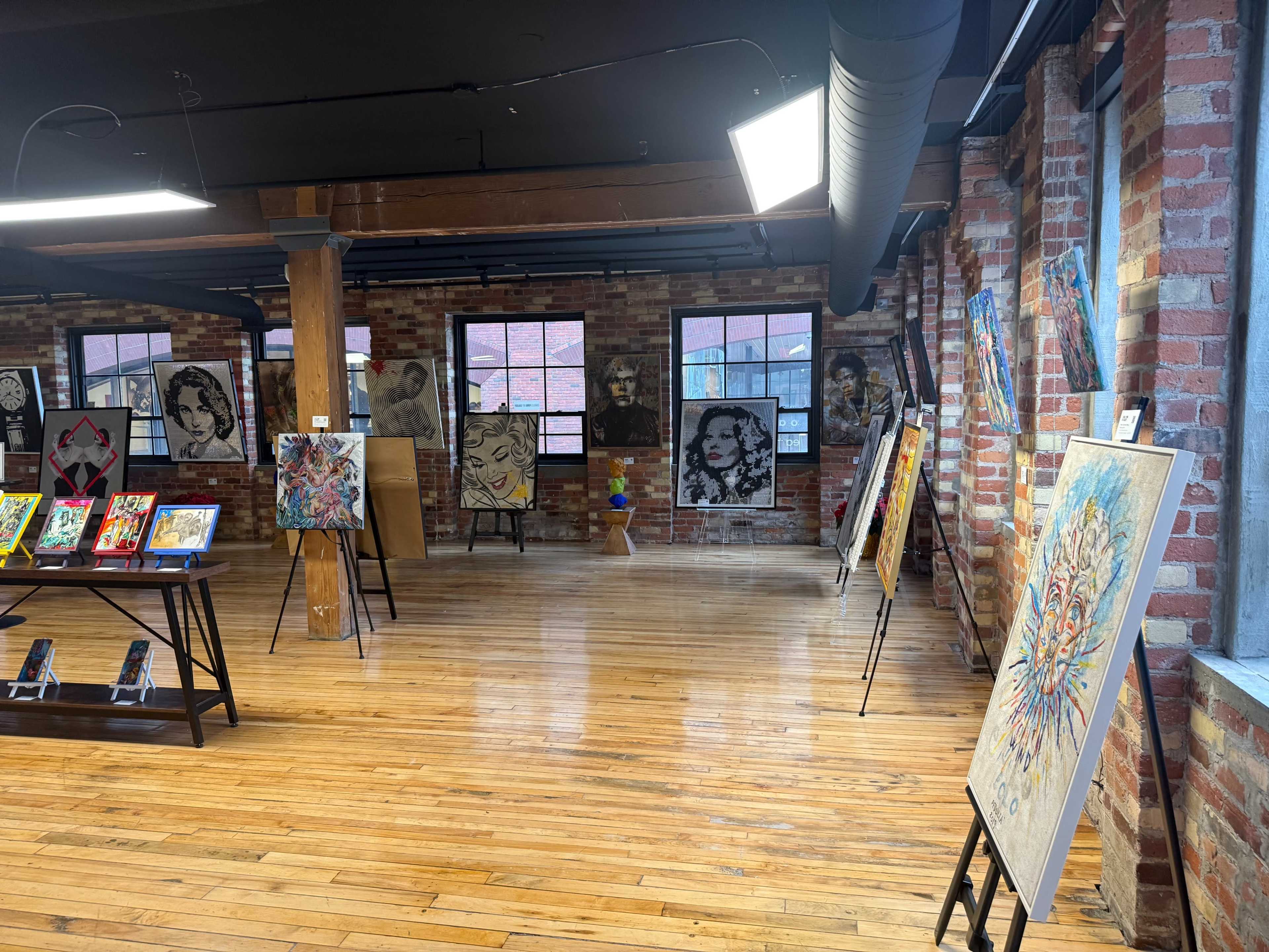 The image shows an art gallery with brick walls, wooden floors, and various paintings displayed on easels and walls.