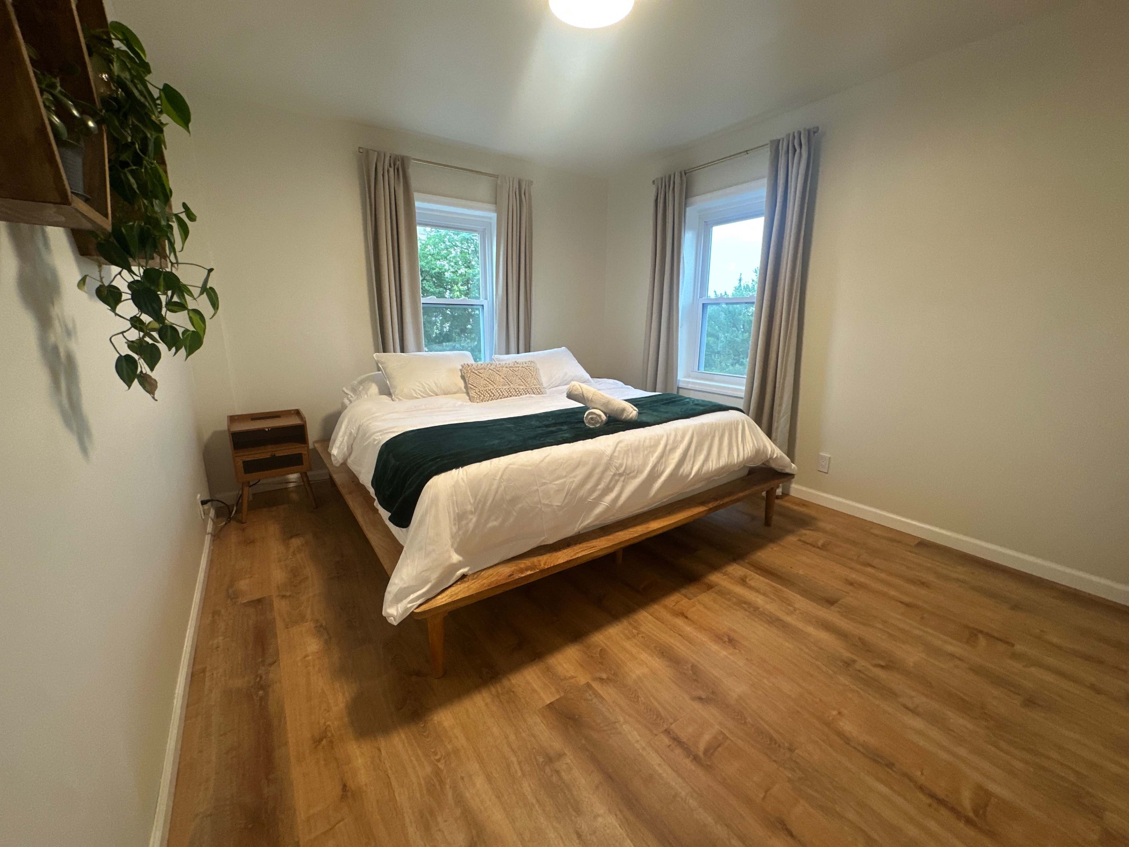 The image shows a simple bedroom featuring a bed with a green blanket and white sheets, two windows, a wooden floor, and a plant on the wall.
