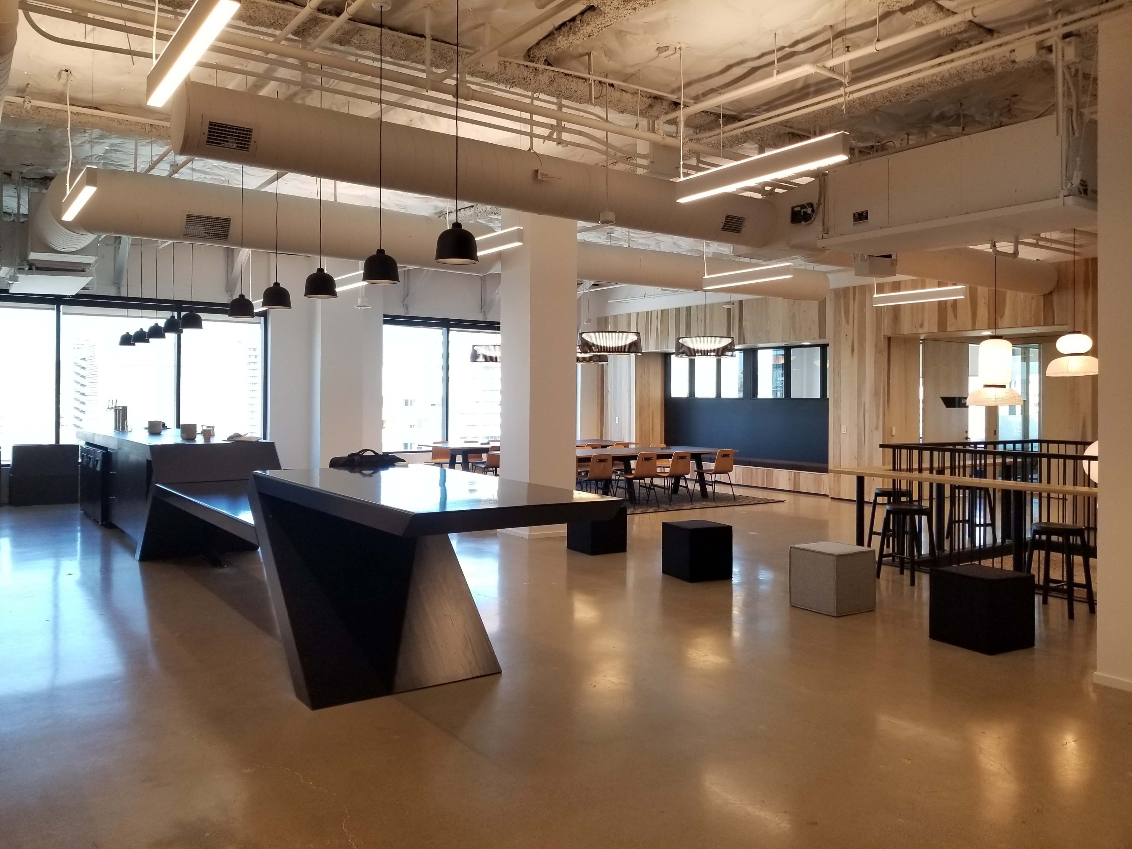 A modern office space featuring a large central table, a kitchen area, and several seating arrangements near windows.