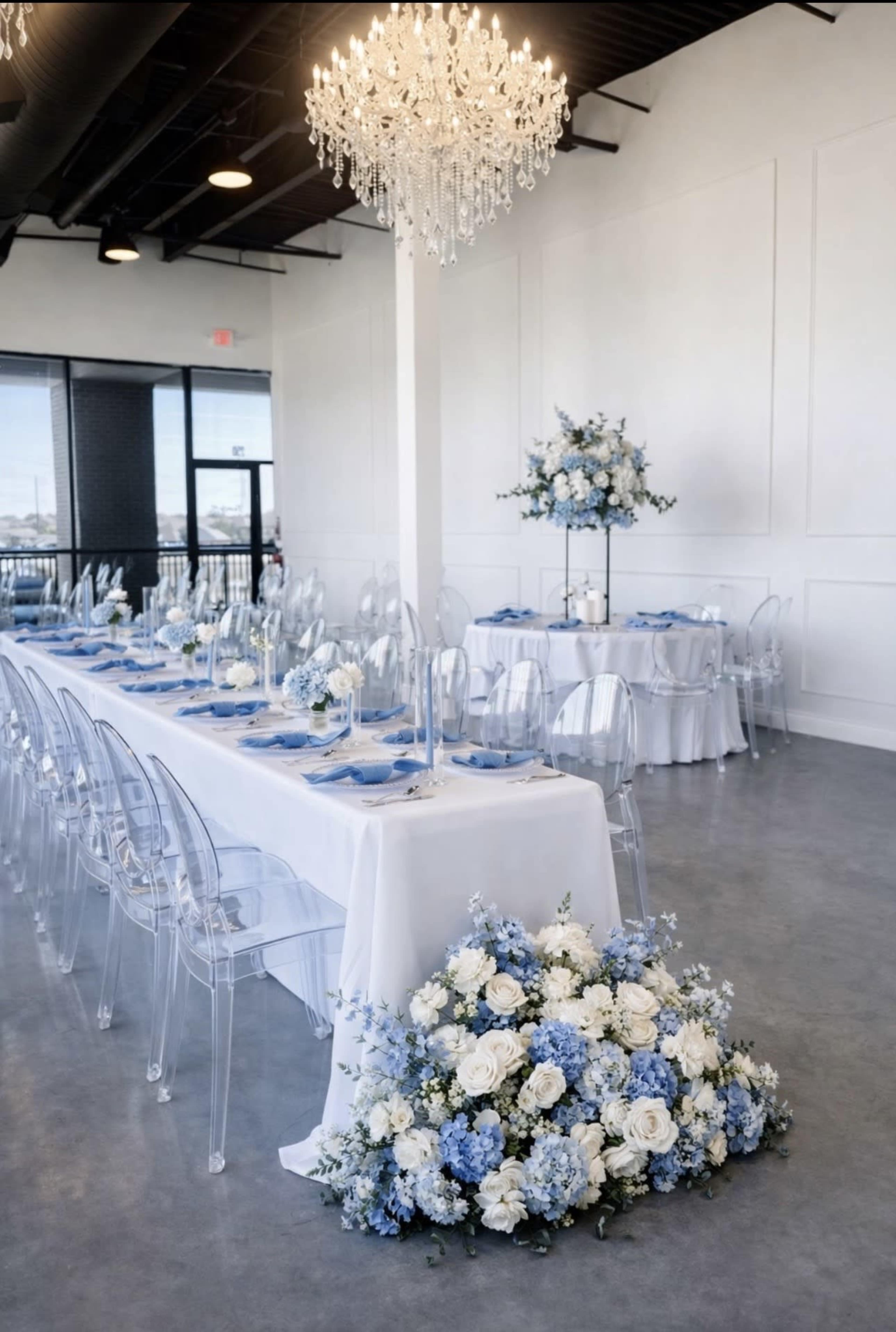 A spacious dining area features a long table set in blue and white decor, adorned with floral arrangements and a crystal chandelier overhead.