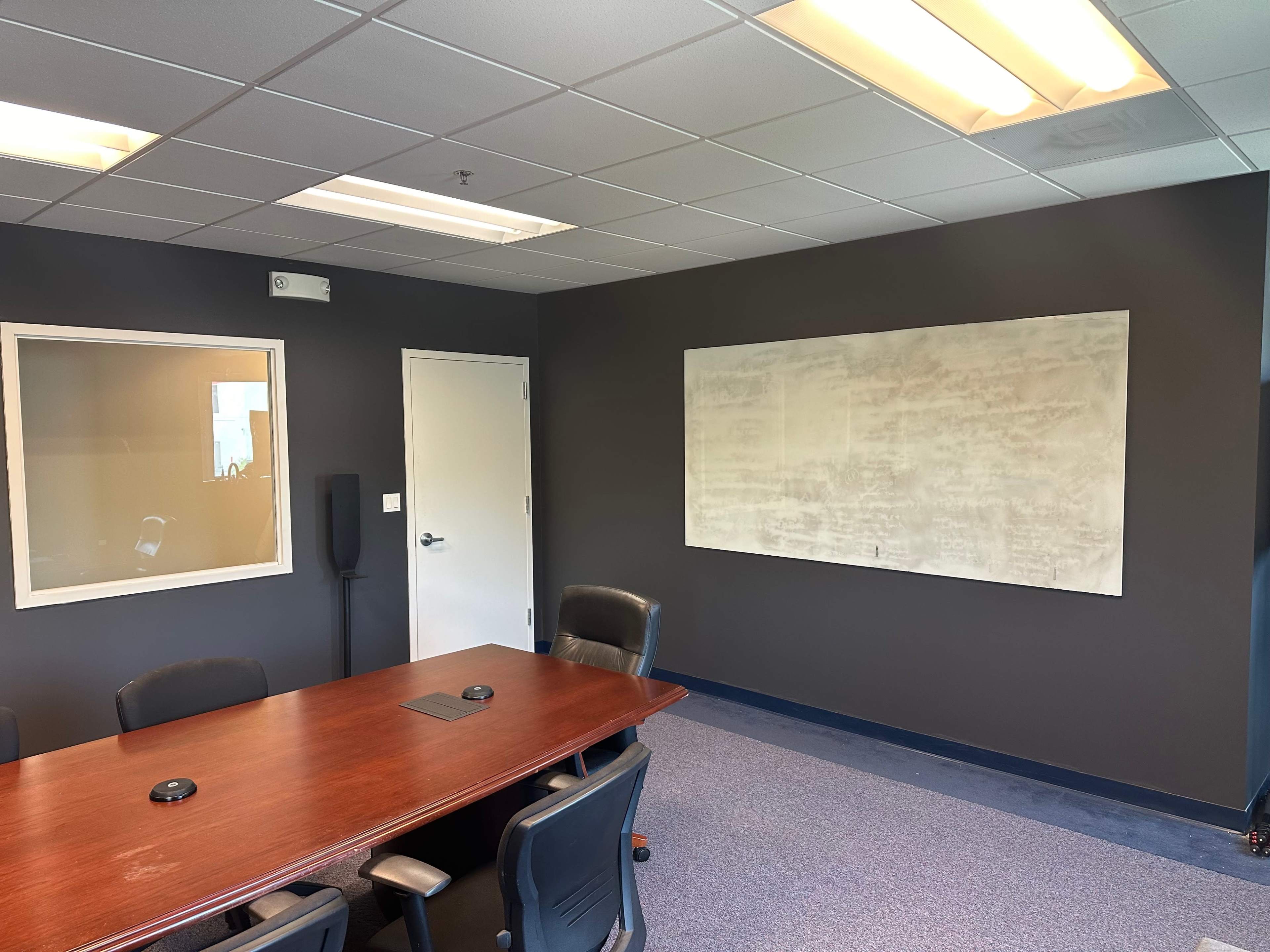 A conference room with a wooden table surrounded by black chairs, a large whiteboard on the wall, and a glass-paneled door.