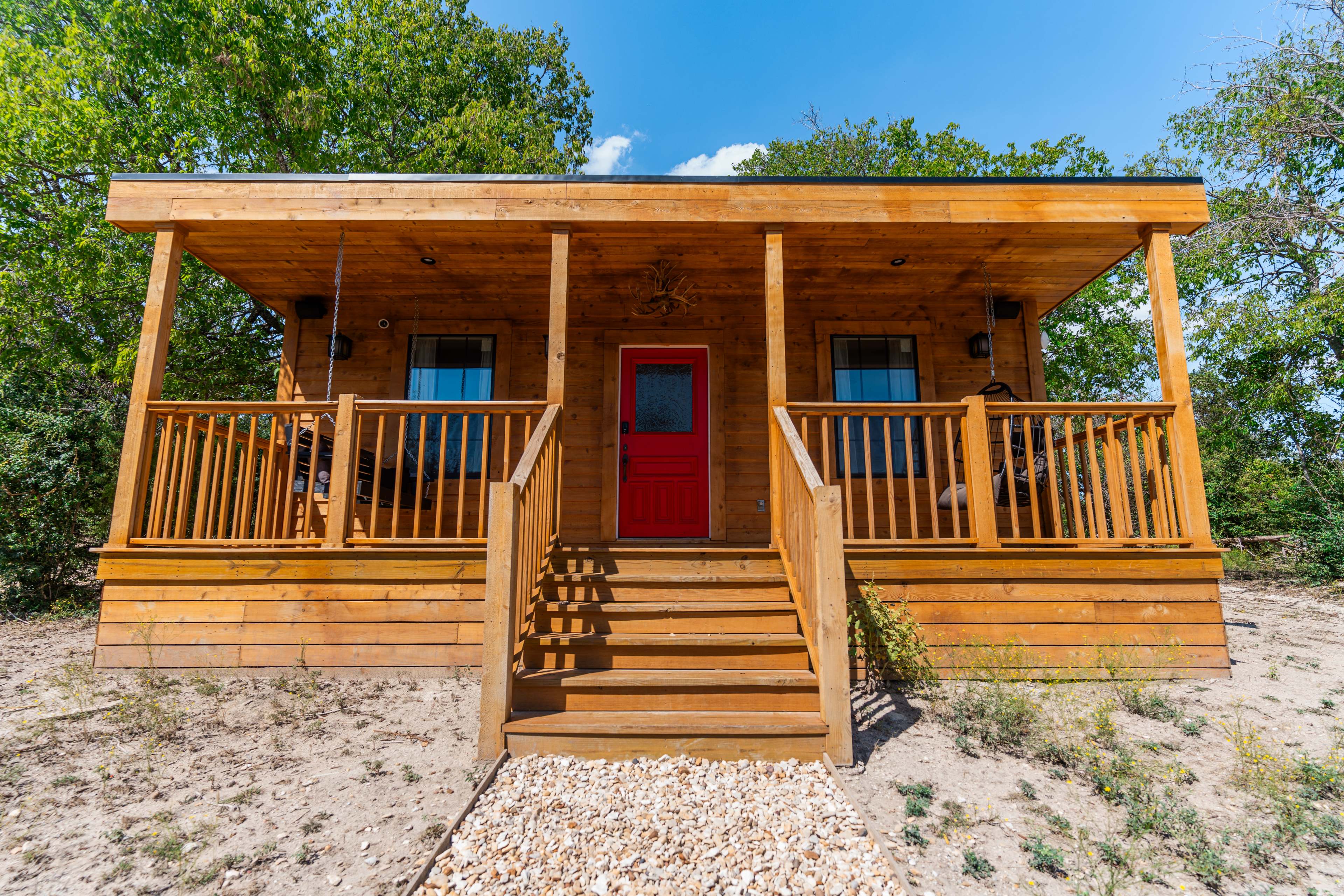 A wooden cabin with a red front door and a porch sits on a sandy area, surrounded by trees.