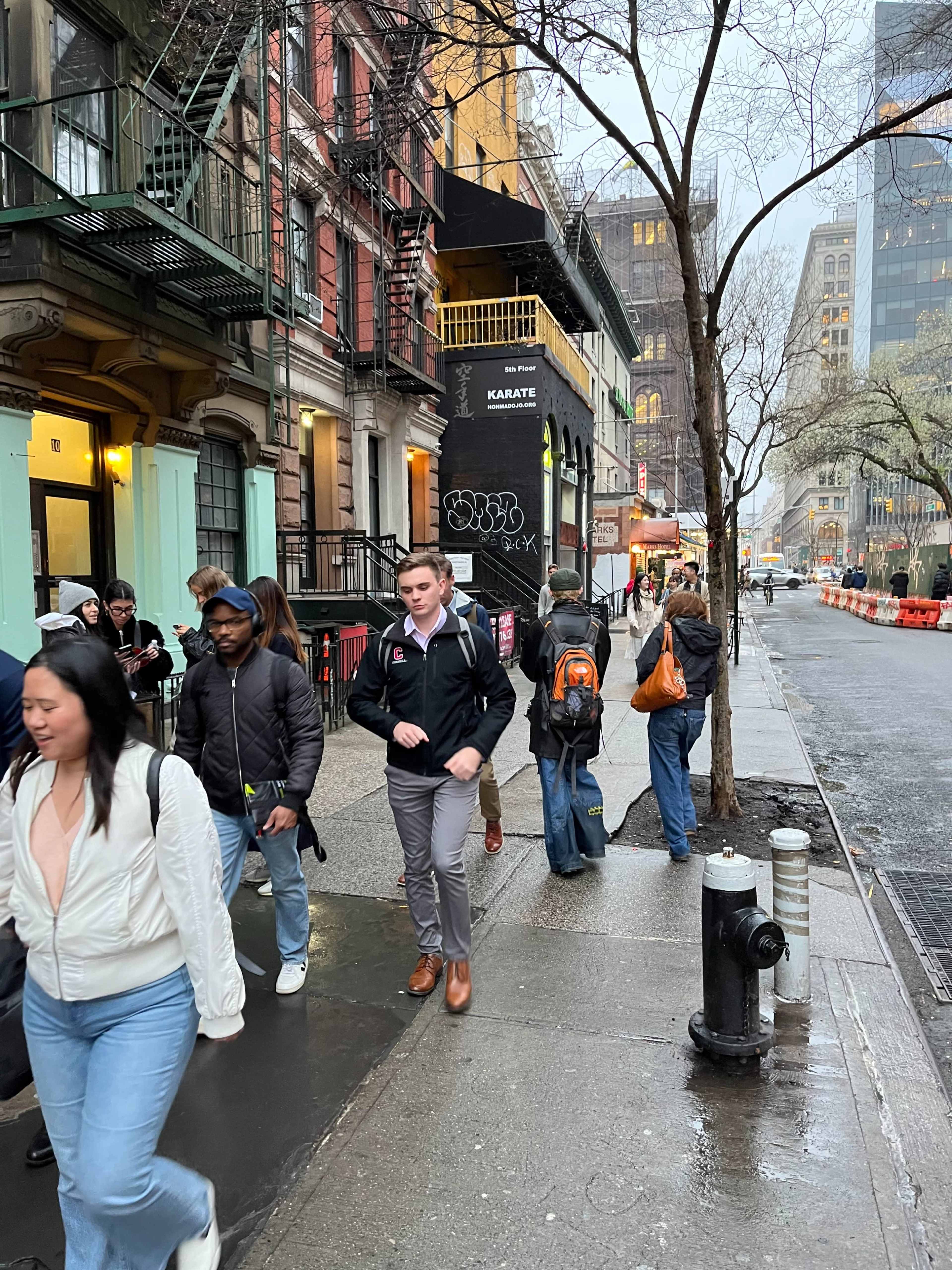 A diverse group of people walks along a rainy street lined with colorful buildings and storefronts.