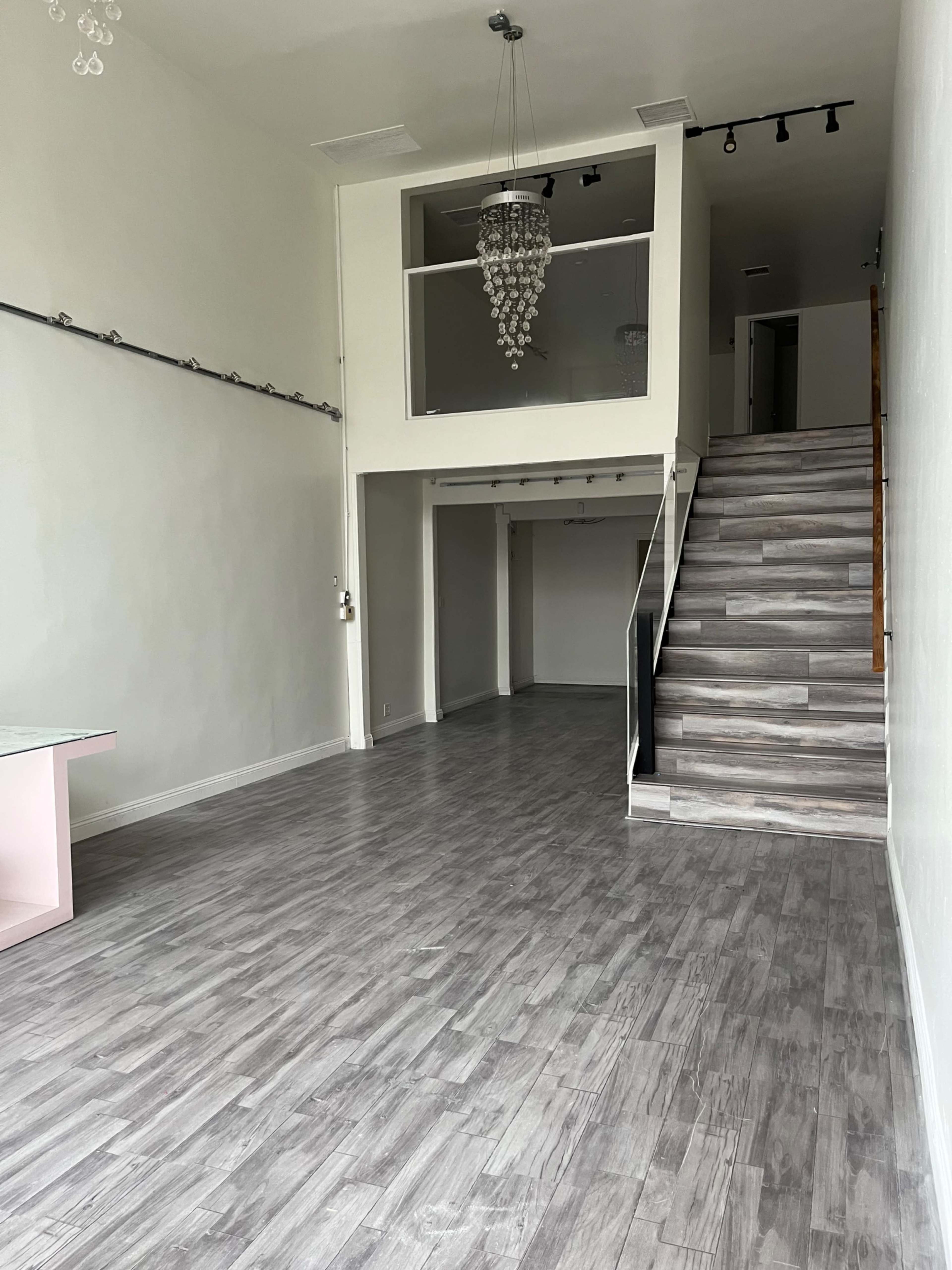 The image shows a spacious room with a wooden staircase leading to an upper level, featuring light-colored walls and a modern chandelier.