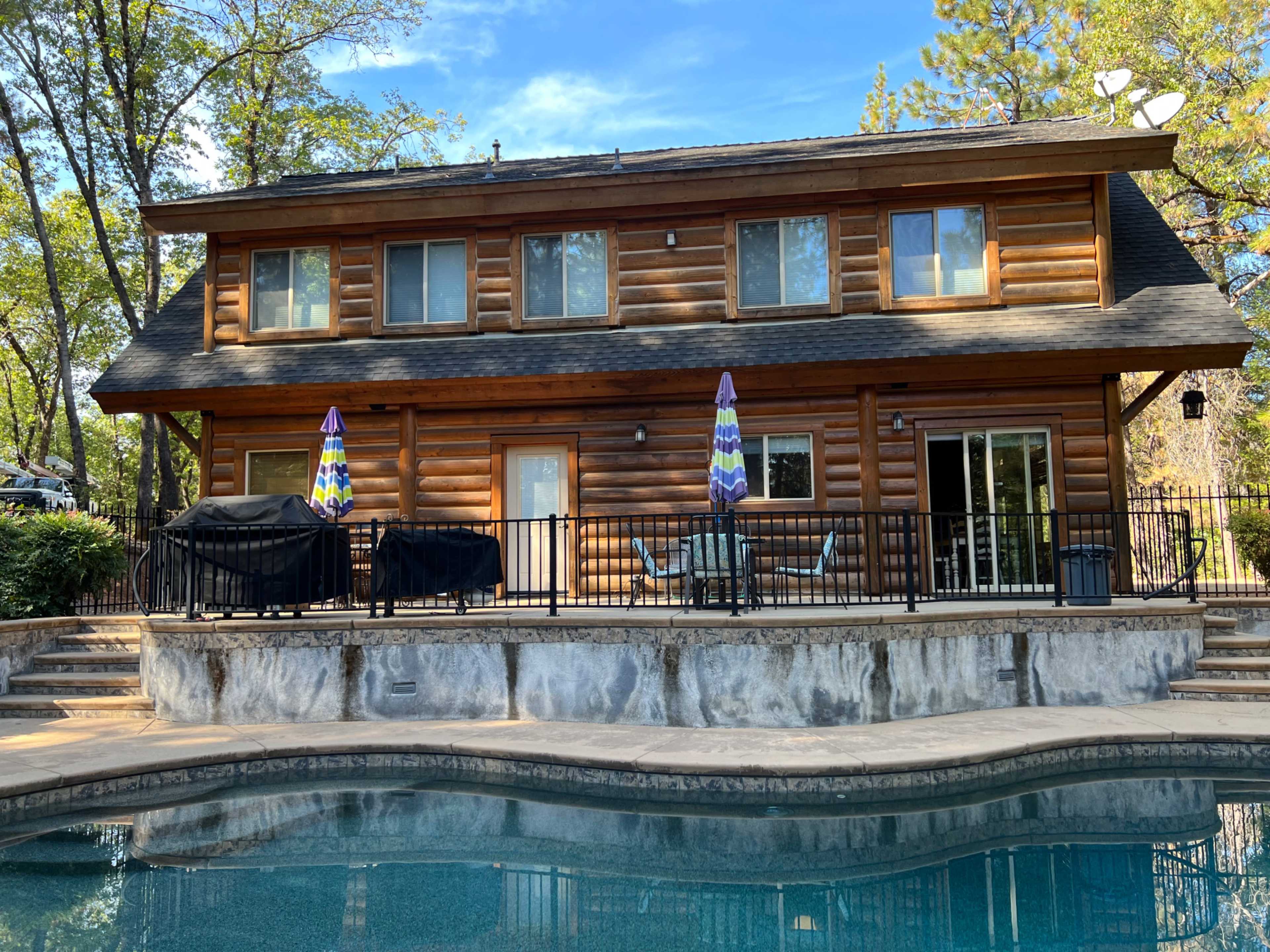 A two-story wooden cabin with a stone patio and pool is surrounded by trees.