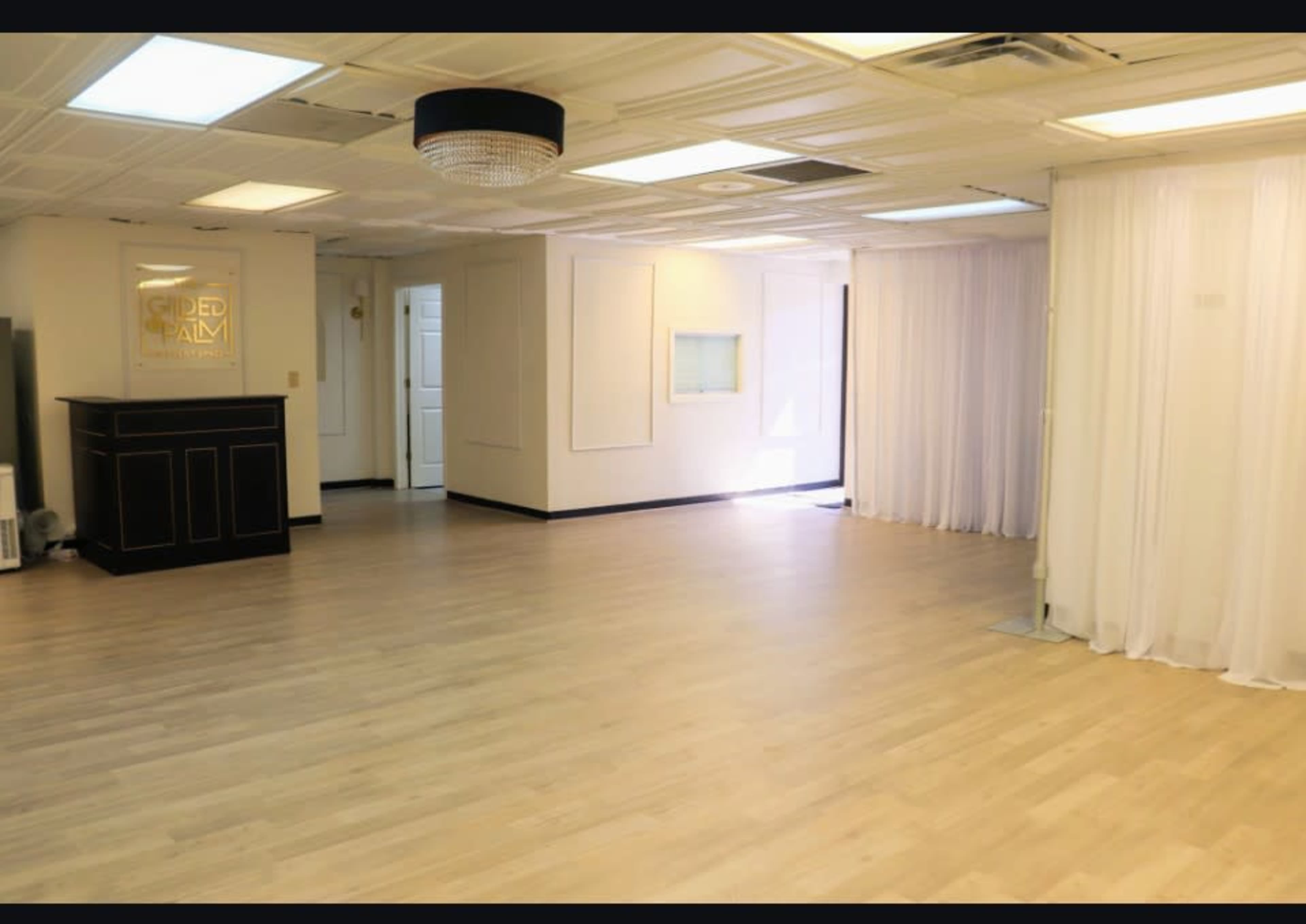 The image shows a spacious, well-lit room with light hardwood flooring, white drapes on one side, and a black reception desk against the wall.