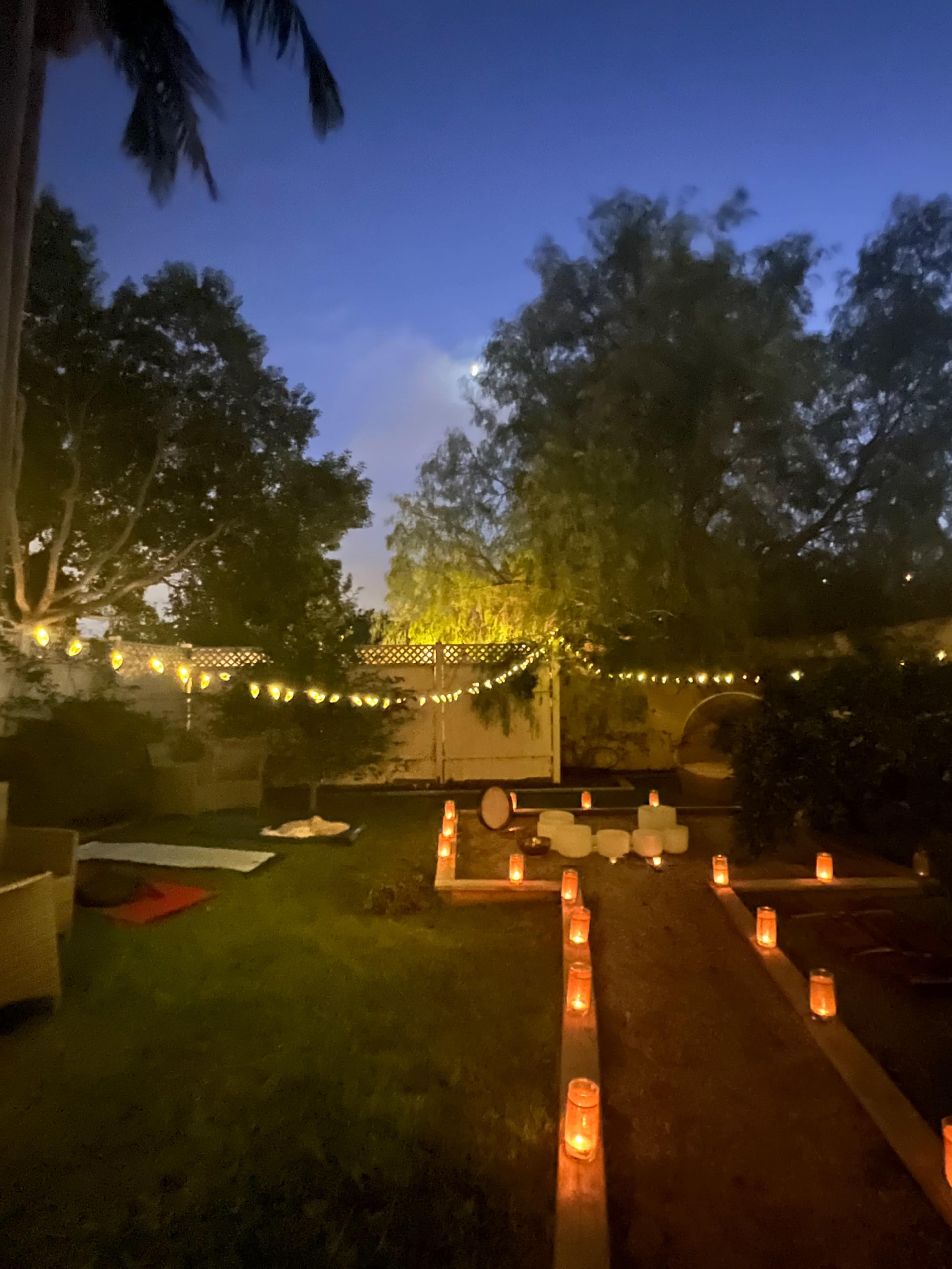 The image shows a dimly lit garden path lined with candles, string lights overhead, and greenery in the background beneath a twilight sky.